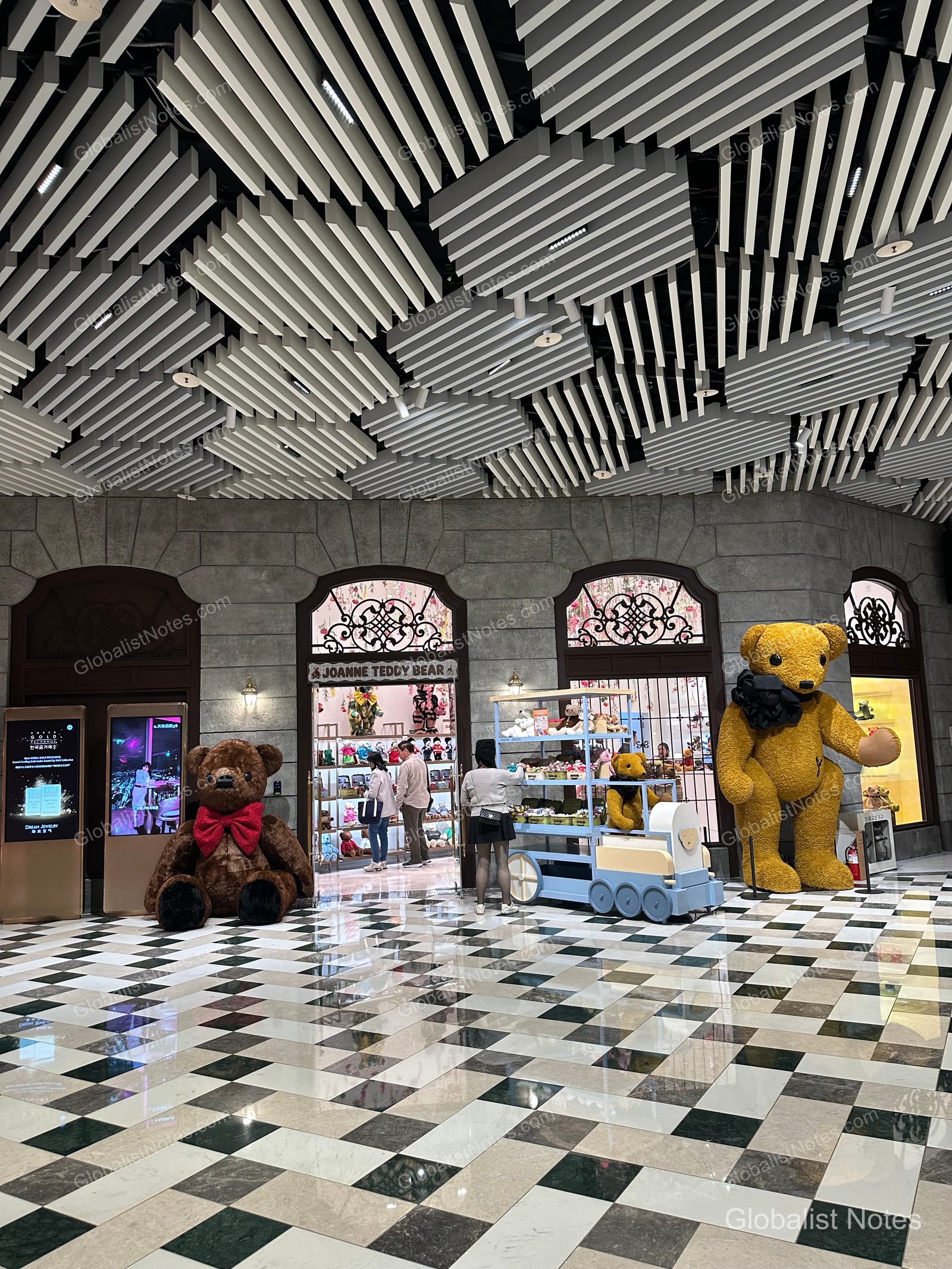 Entrance to the Joanne Teddy Bear shop at Grand Hyatt Jeju. Large brown and yellow teddy bear statues stand on either side of the entrance, set against a black-and-white checkered marble floor. The ceiling features a geometric grid structure.
