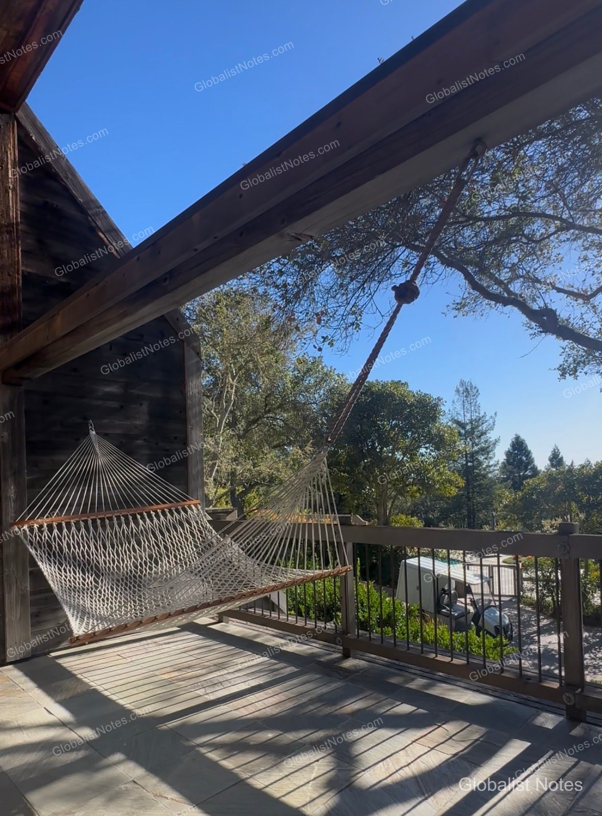 The private balcony of the Alila Ventana Big Sur Junior Suite featuring a white mesh hammock on a wooden deck, with a view of trees, the sky, and a golf cart parked below.