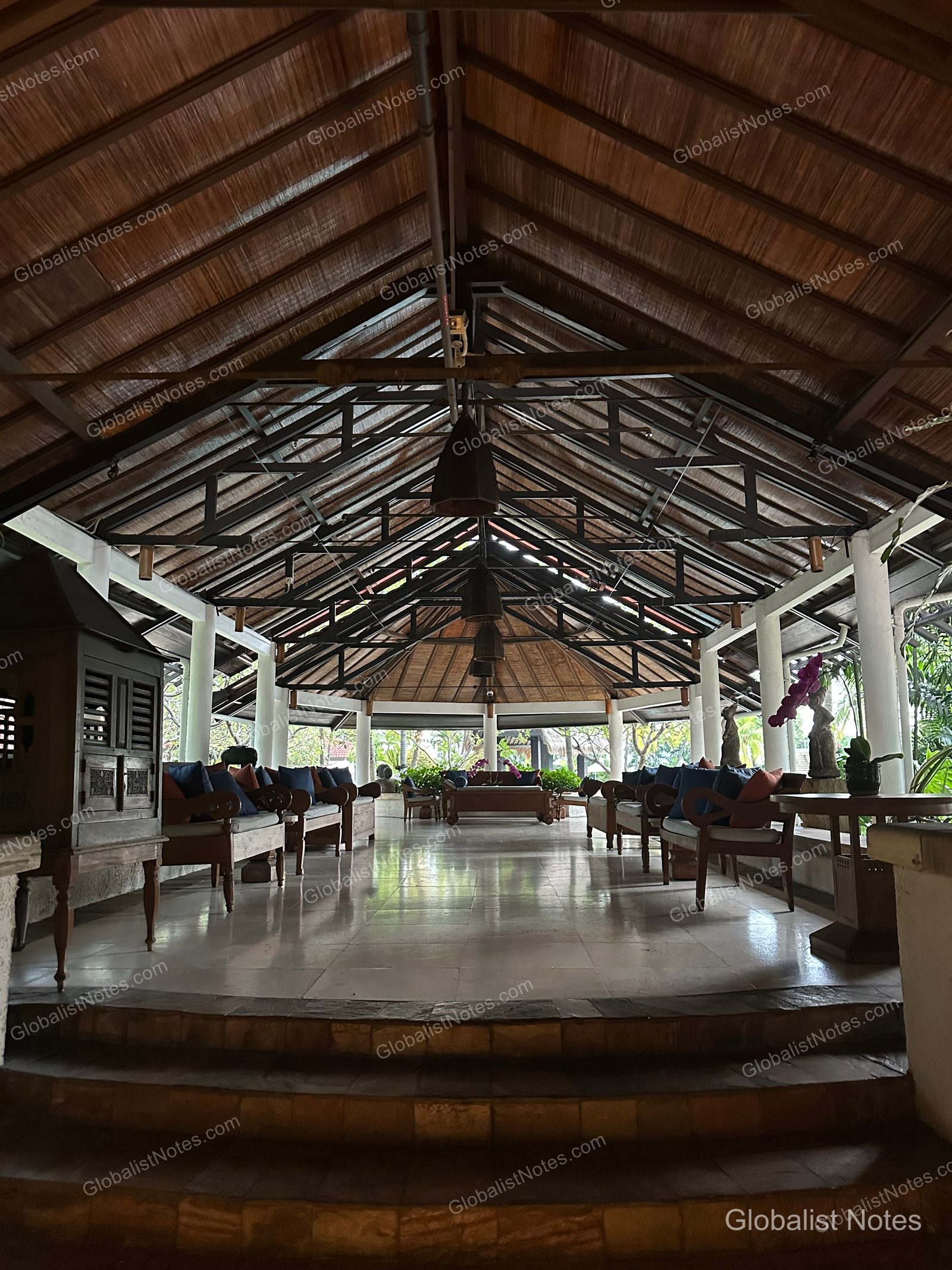 The open-air lobby area at Grand Hyatt Jakarta, featuring a high gabled roof with symmetrical wooden frames and columns, tiled floors, comfortable sofas and chairs, and an open layout overlooking the garden.
