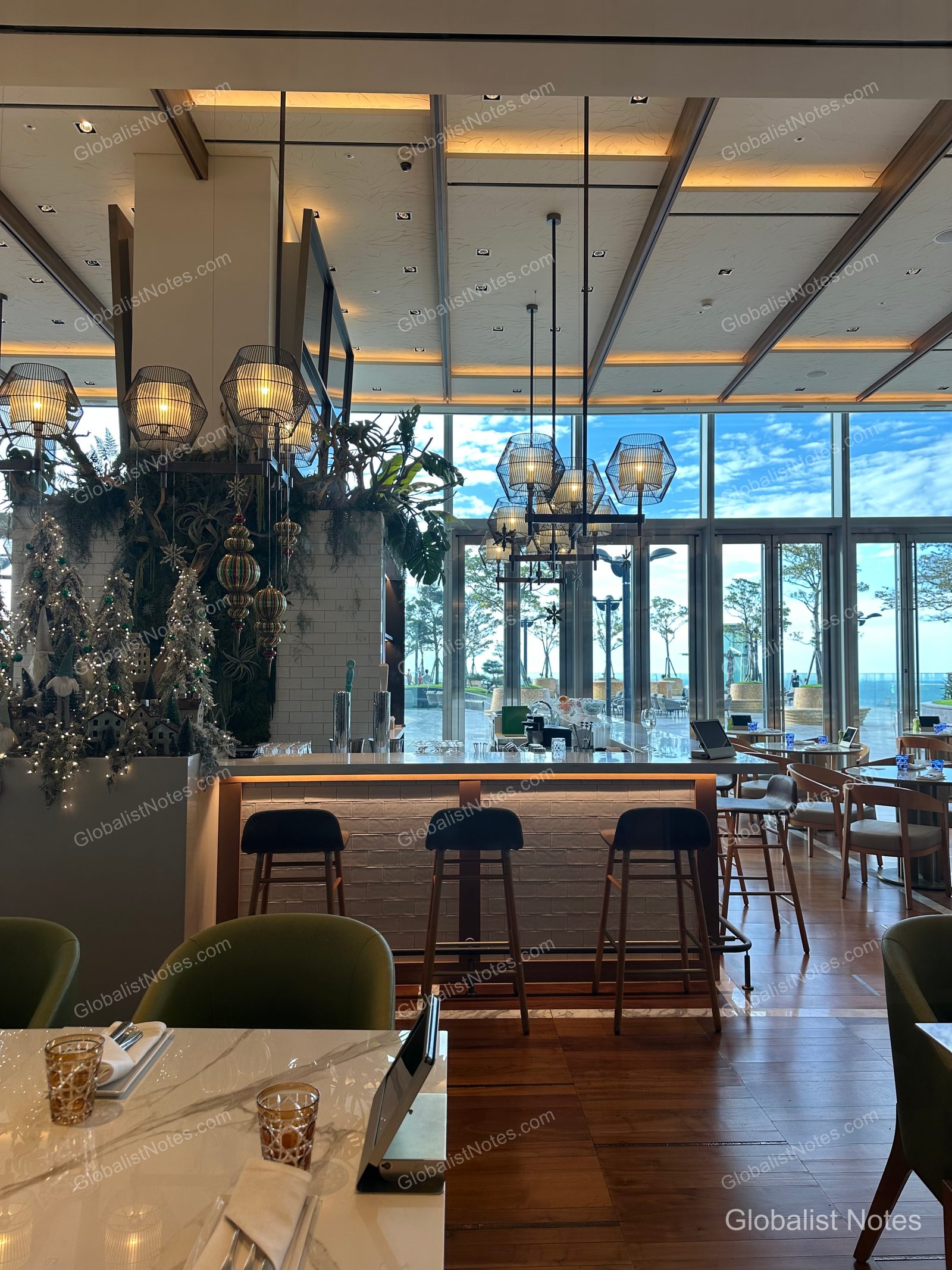The bar area at Grand Hyatt Jeju, featuring high ceilings with geometric pendant lighting and floor-to-ceiling windows overlooking the Jeju sky. High chairs are arranged at the bar, and Christmas trees and decorations are set up, creating a calm and sophisticated atmosphere.