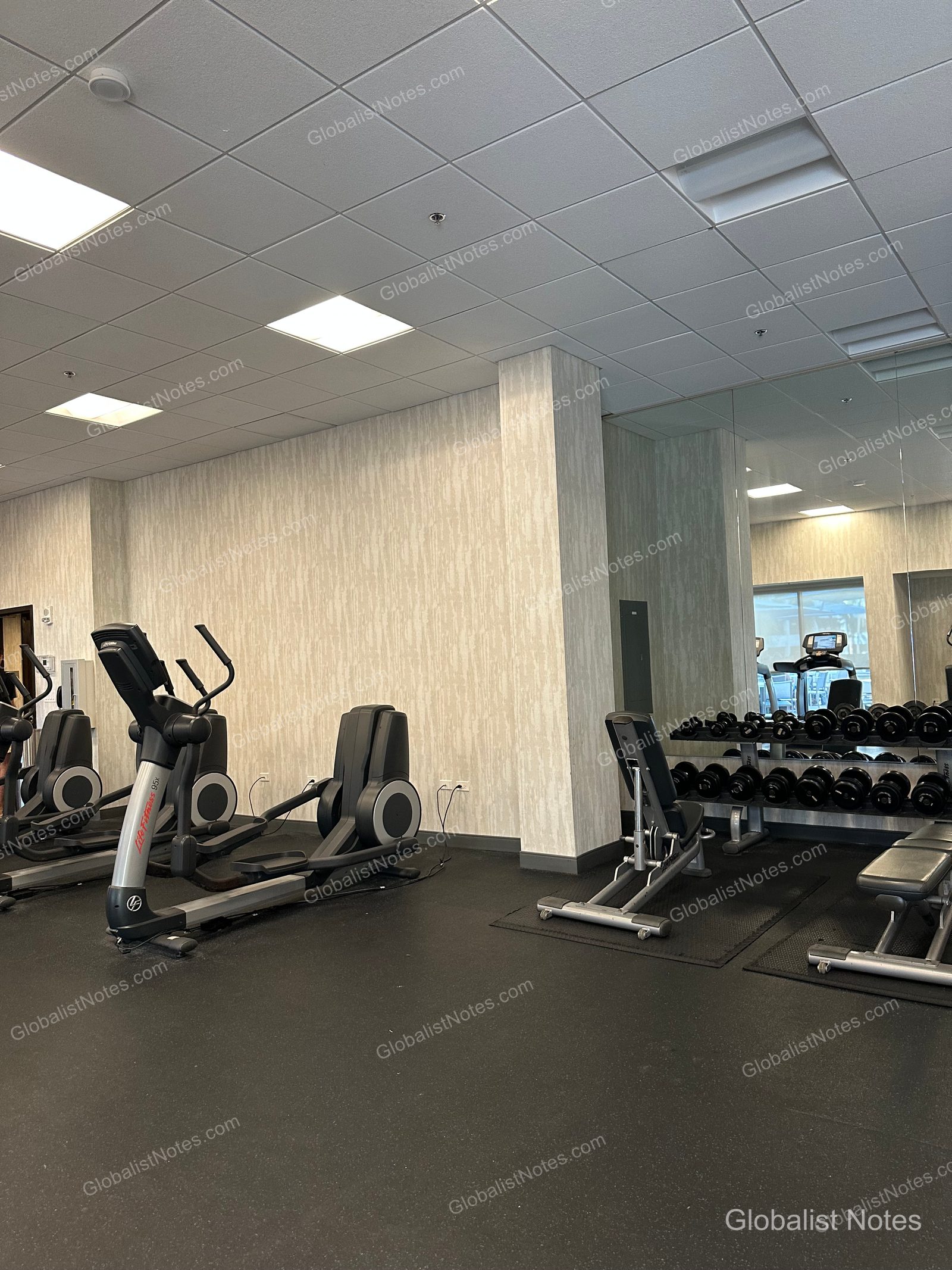 Fitness center at Hyatt Place Waikiki Beach in Honolulu, Hawaii, featuring elliptical machines, a dumbbell rack, and a workout bench on a black rubber floor, with a mirrored wall that makes the space feel larger.