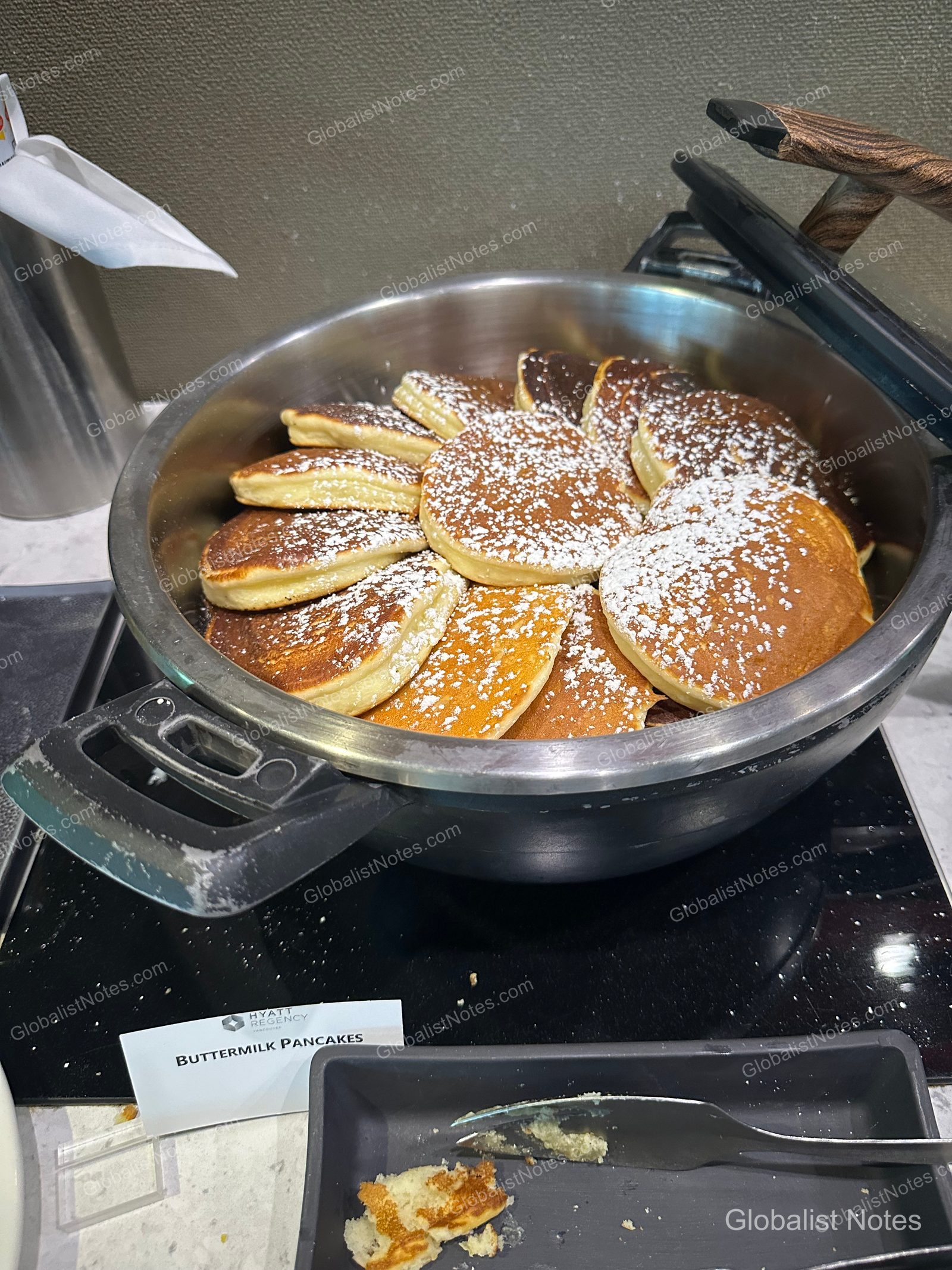 Buttermilk pancakes dusted with powdered sugar inside a stainless steel warmer at the Hyatt Regency Vancouver breakfast buffet, with a menu label reading 'BUTTERMILK PANCAKES' and serving tongs placed in front.