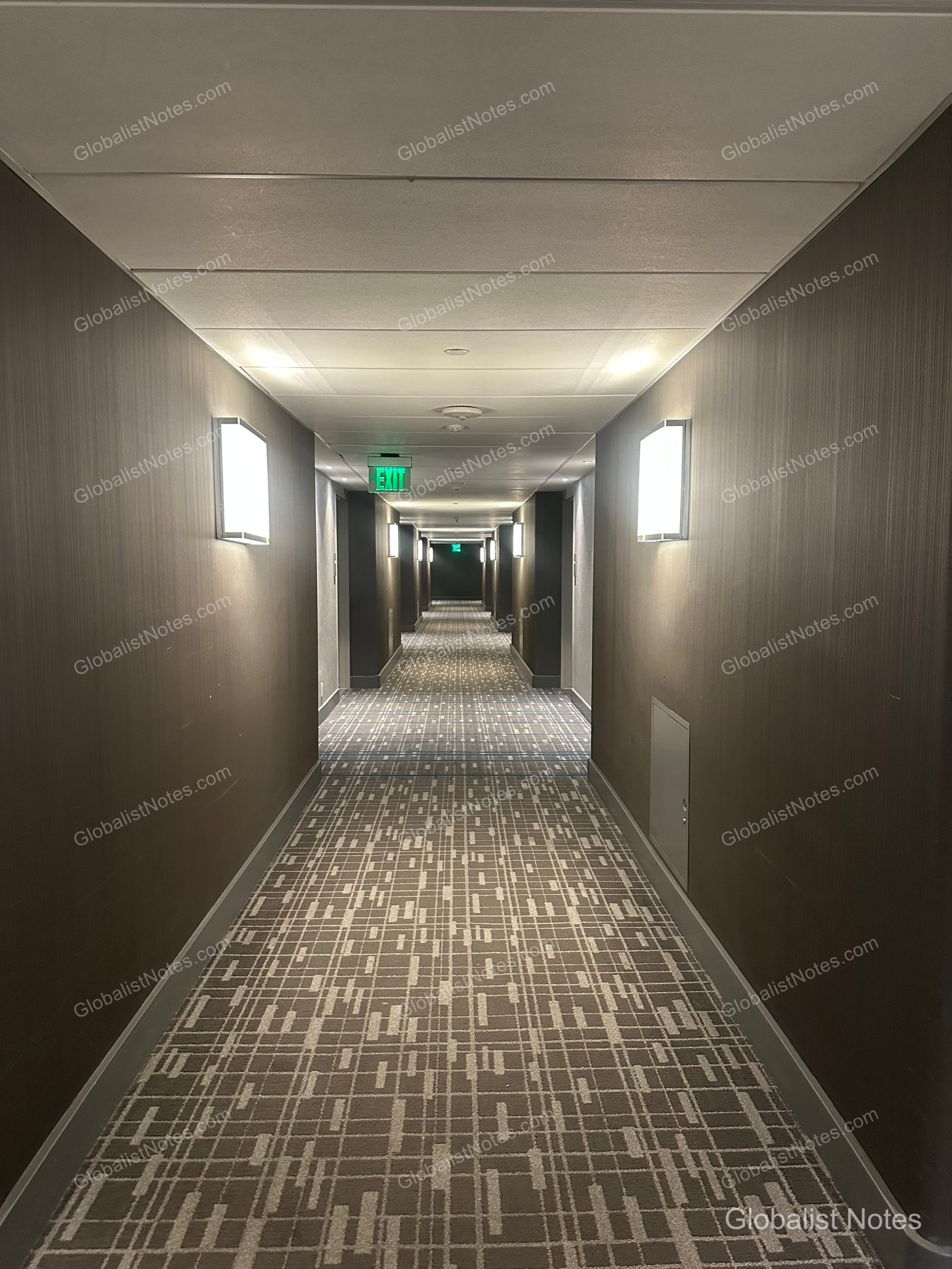 Guest room hallway at Hyatt Regency Los Angeles International Airport. The long corridor features dark walls, geometric-patterned carpeting, wall-mounted sconces on both sides, and an illuminated green exit sign at the far end.