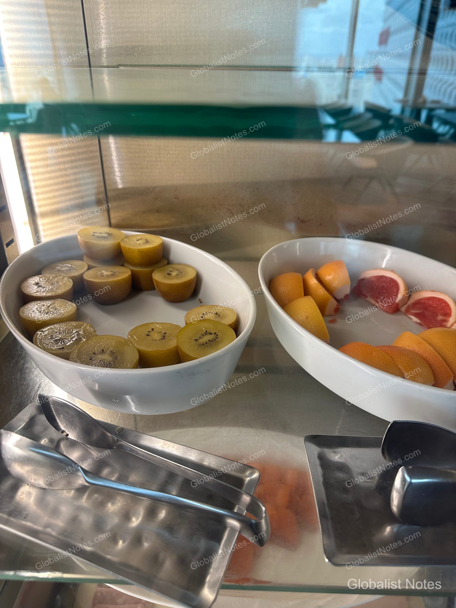 A view of the breakfast buffet showcase at Grand Hyatt Jeju, featuring two white bowls side-by-side. The left bowl contains prepared gold kiwi, and the right bowl holds sliced grapefruit and oranges, each with a metal serving tong placed in front.