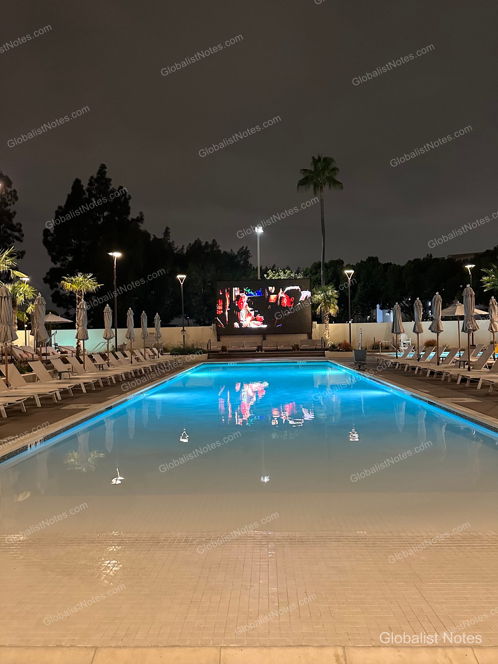 Hyatt Regency Irvine 夜間戶外泳池全景。泳池末端的大螢幕正播放影像，兩側整齊排列著躺椅與遮陽傘，燈光氛圍溫馨。