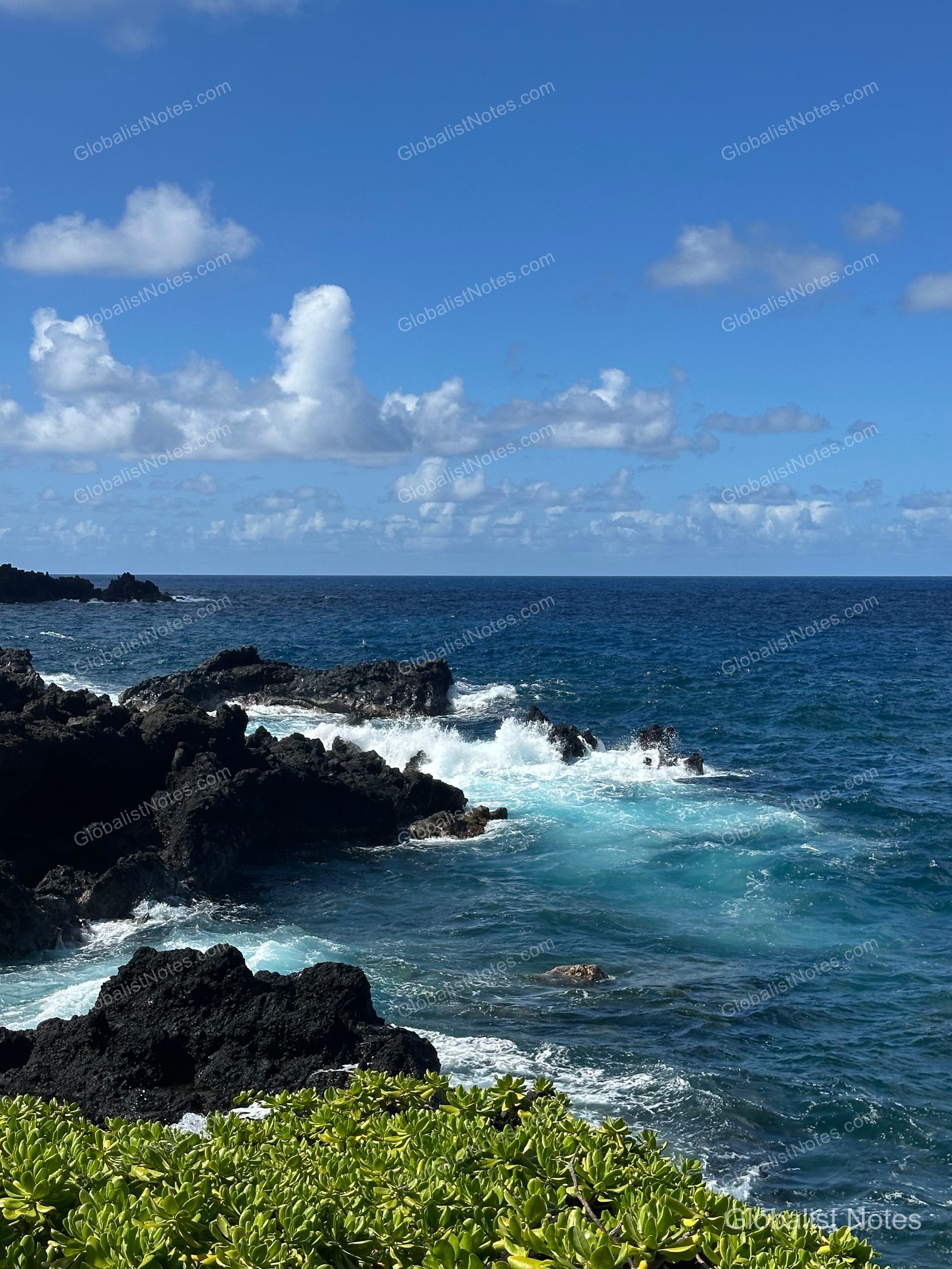 Coastal view near Hana-Maui Resort, featuring blue waves crashing against black volcanic rocks with the horizon and clear sky in the distance.