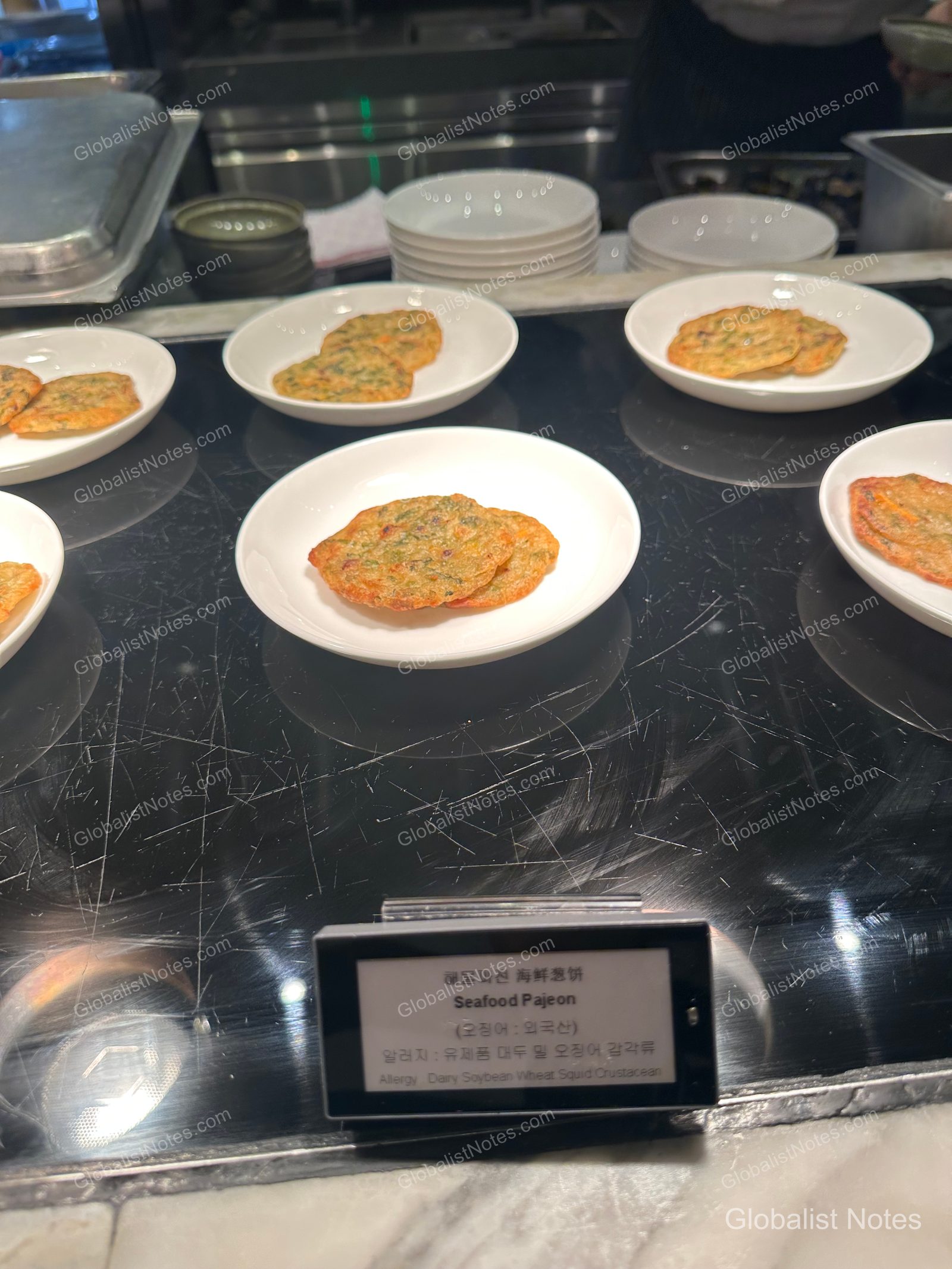 White plates of seafood pajeon (scallion pancakes) displayed on a warming showcase at the Grand Hyatt Jeju lounge buffet, with a sign in front detailing the dish name and allergen information.