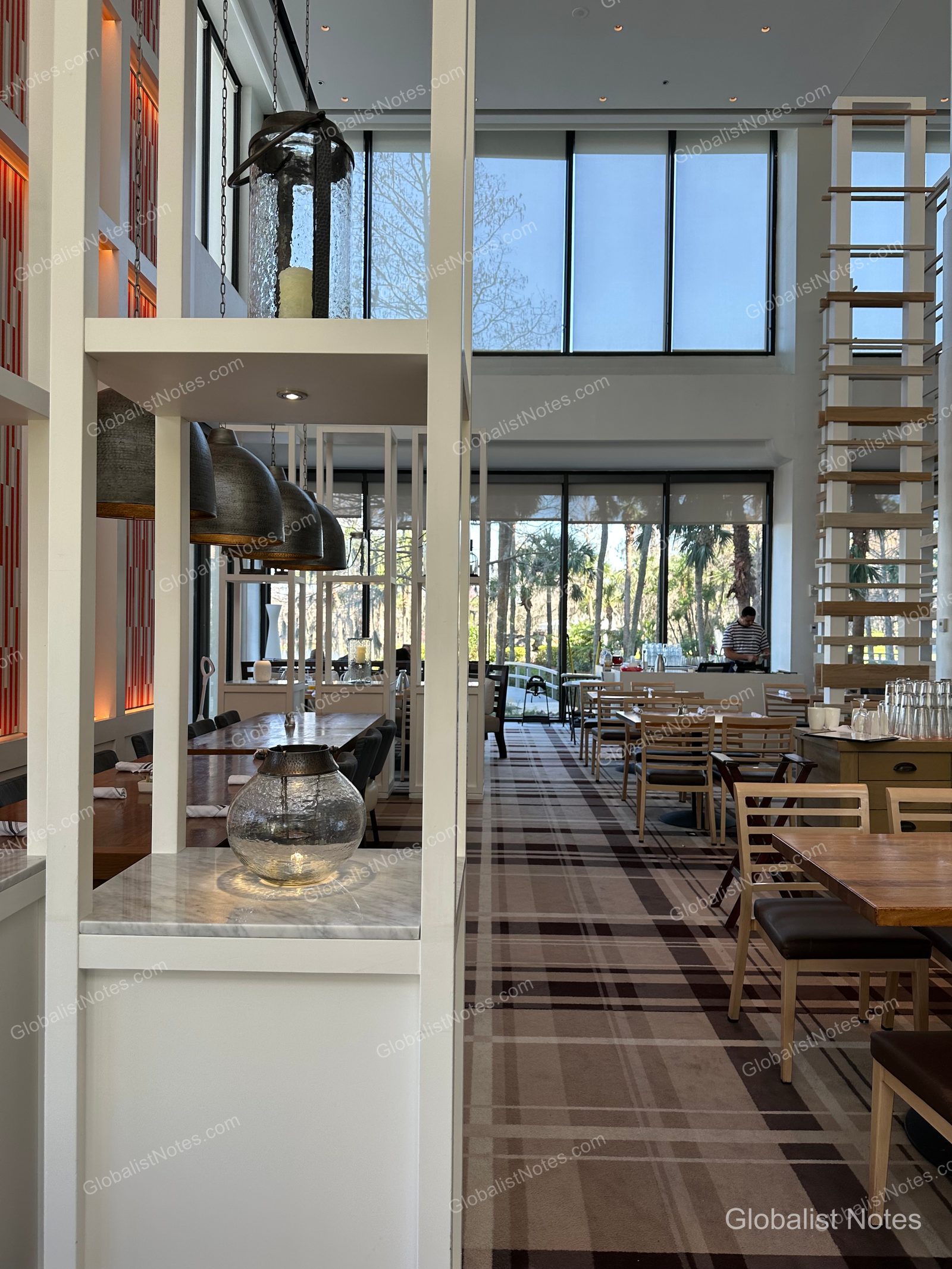 High-ceilinged dining area at Hyatt Regency Grand Cypress. Wooden chairs and tables are arranged on a patterned carpet, with large floor-to-ceiling windows looking out onto the garden and trees.