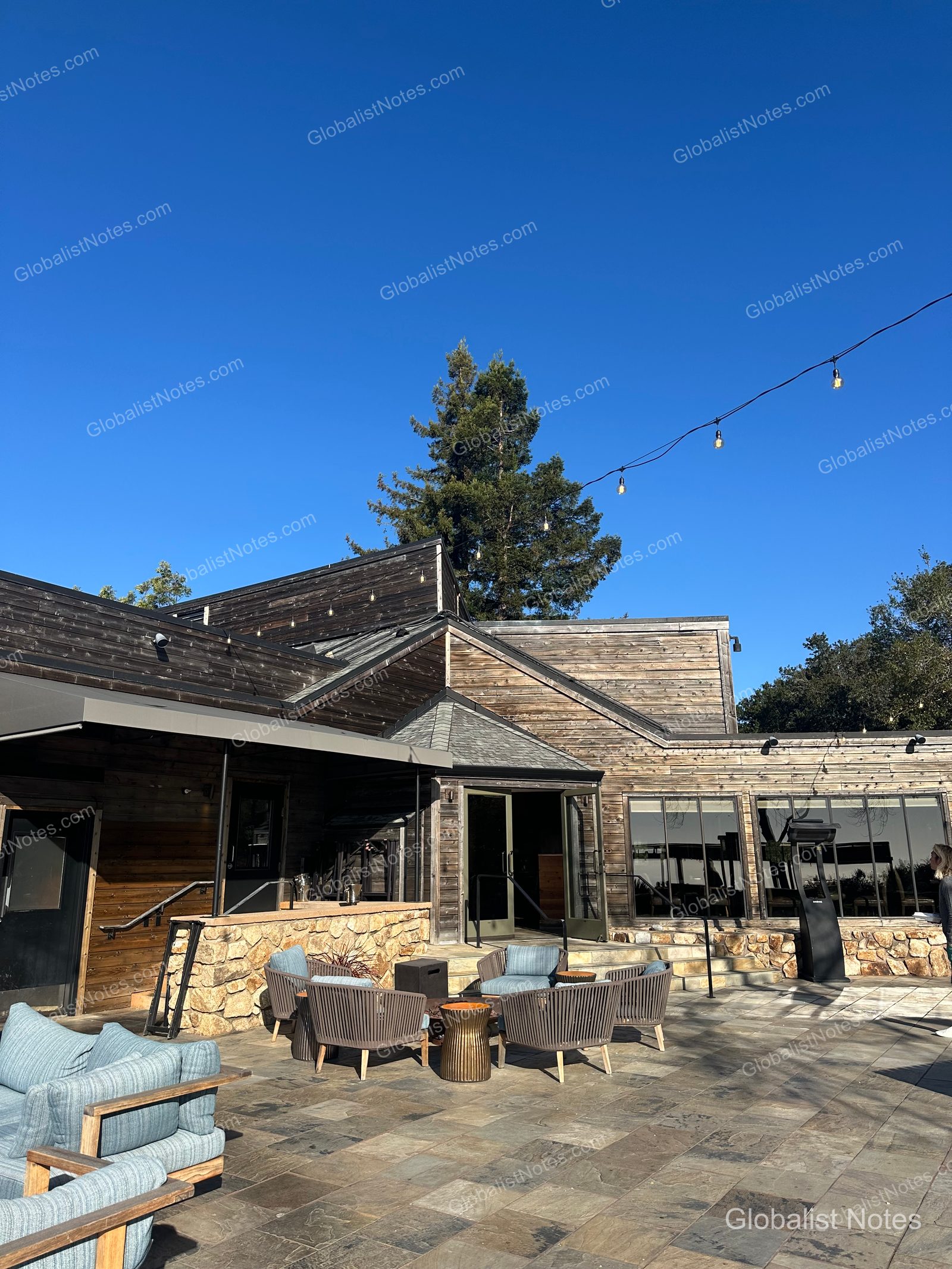The outdoor terrace at Alila Ventana Big Sur under a clear blue sky. Outdoor chairs with blue cushions and tables are arranged on a stone floor in front of a dark brown wooden building, with string lights hanging overhead.