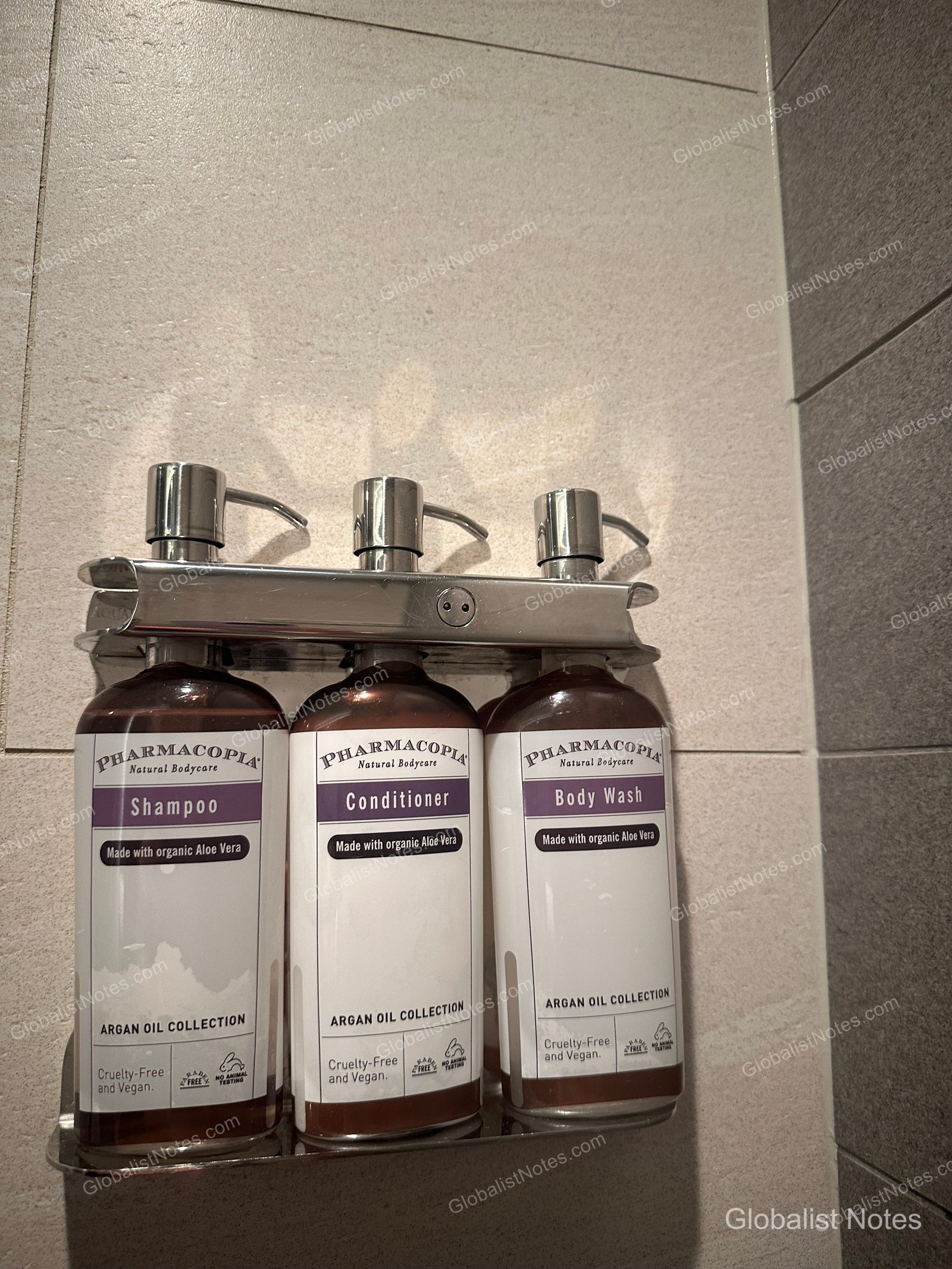 Three pump bottles of Pharmacopia Argan Oil collection shampoo, conditioner, and body wash mounted on a metal rack on the bathroom wall at Hyatt Regency Seattle. Each bottle is labeled as vegan and containing organic aloe vera.