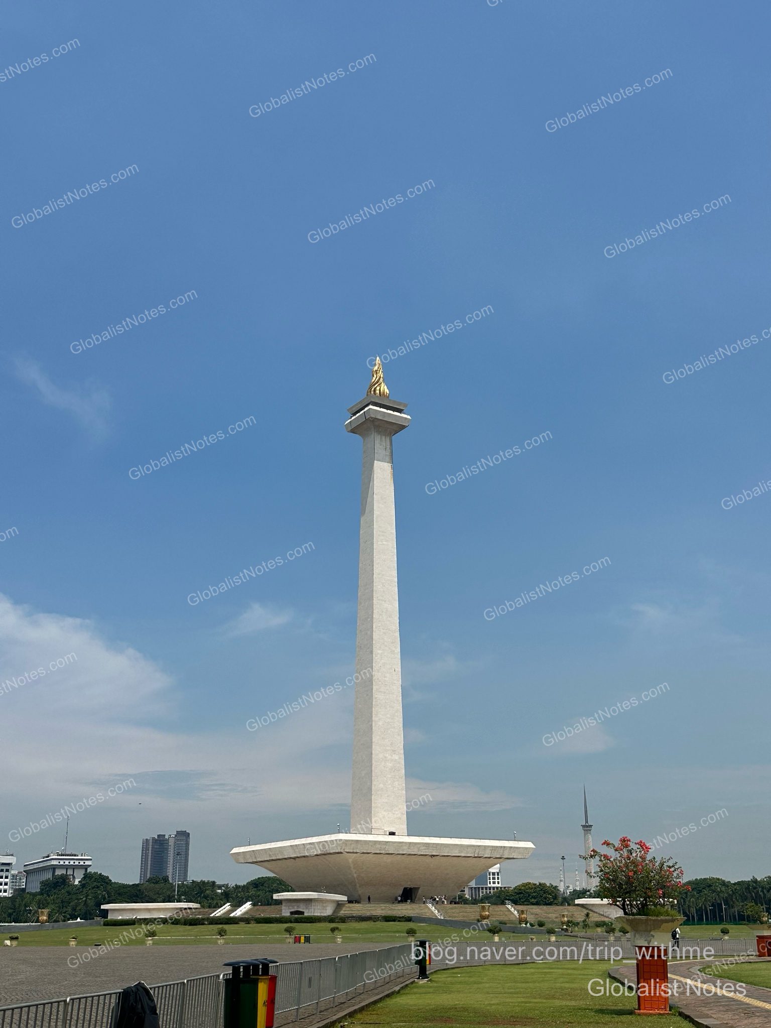 The Monas (National Monument), a symbol of Jakarta, Indonesia, stands tall under a clear blue sky, with its golden flame top and the surrounding grassy plaza clearly visible.