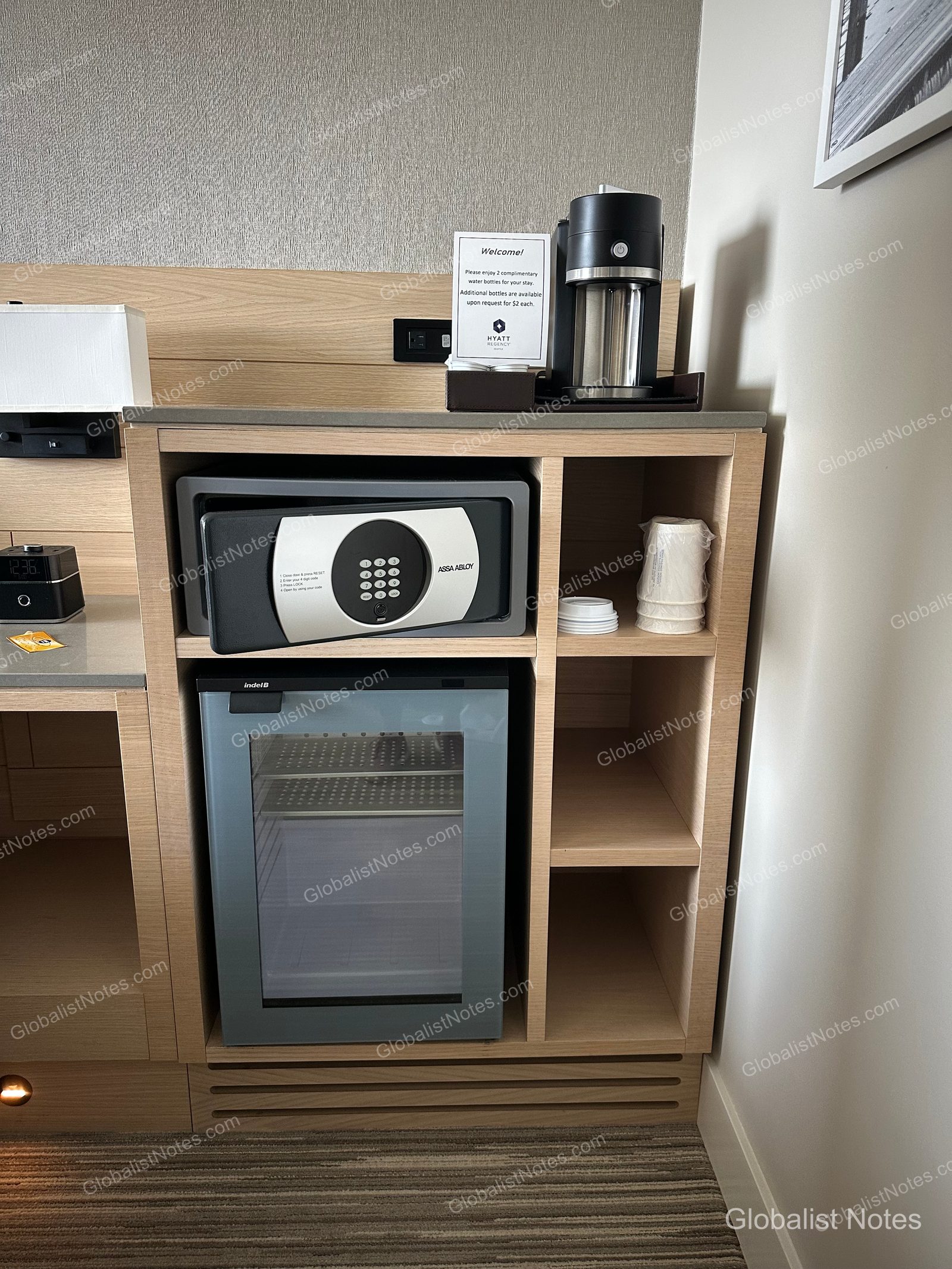 A coffee machine and information card sit on top of the wooden furniture in a Hyatt Regency Seattle Standard King room, with an open safe in the middle shelf and an empty mini-fridge with a glass door and disposable cups stored in the bottom section.