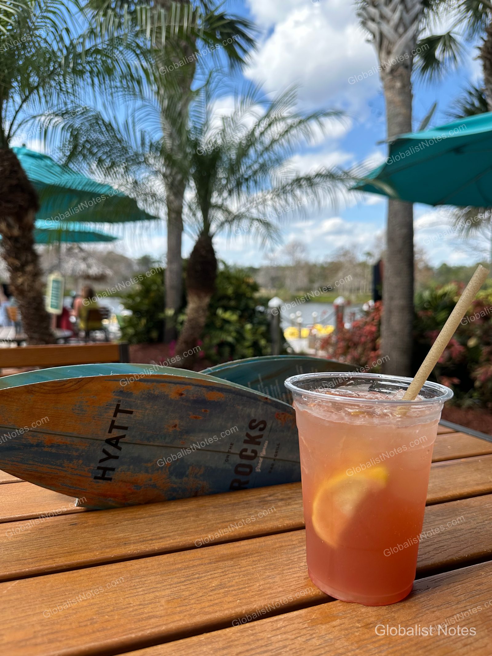 A pink drink on an outdoor table at Hyatt Regency Grand Cypress, with palm trees, blue umbrellas, and a lake view in the background.