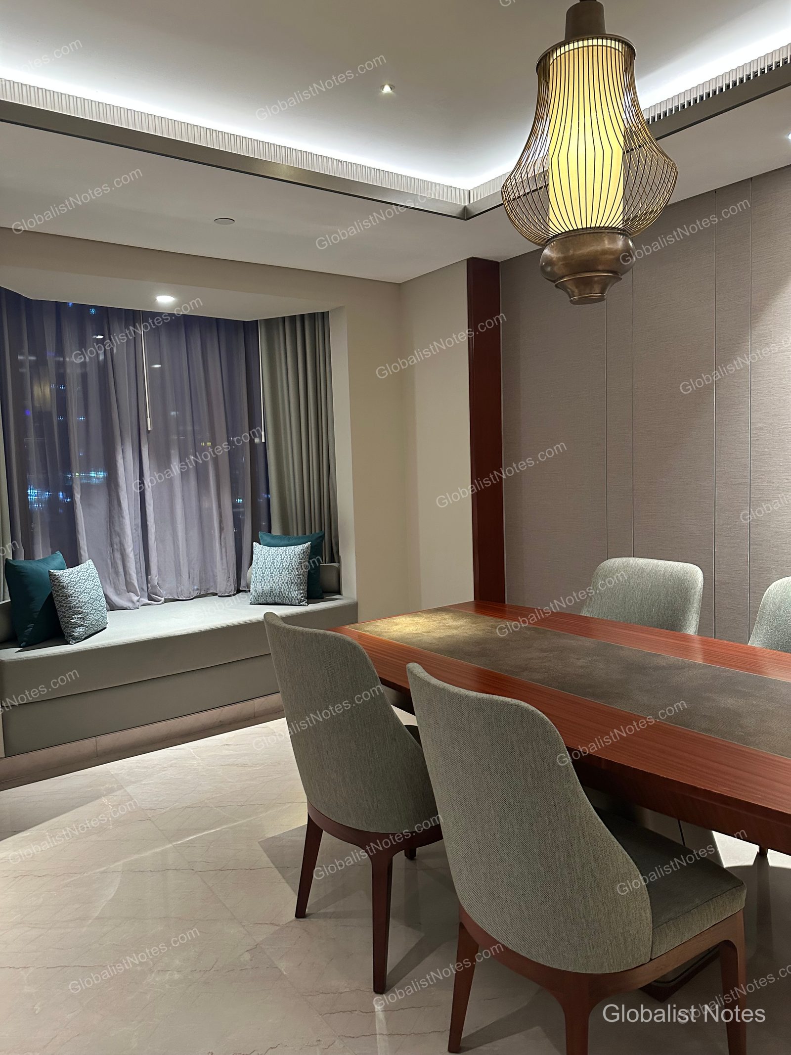 The dining area of a Grand Suite at Grand Hyatt Jakarta, featuring a long wooden table, gray chairs, a unique pendant light, and bench-style seating by the window.