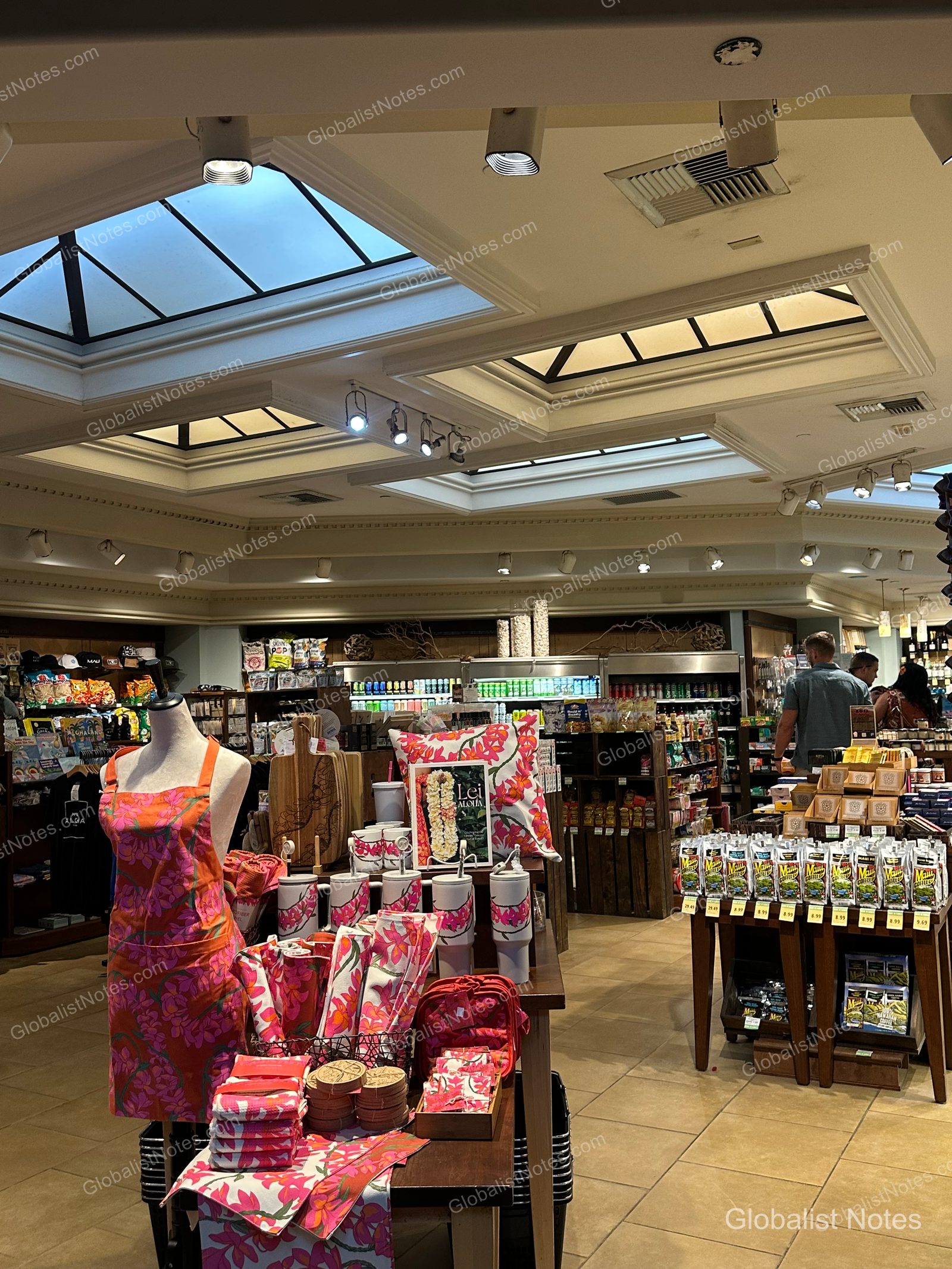 View of the gift shop inside Hyatt Regency Maui Resort and Spa. The shop features a skylight, with floral-print aprons, cushions, and tumblers displayed on central tables, and beverage refrigerators and coffee products visible in the back.