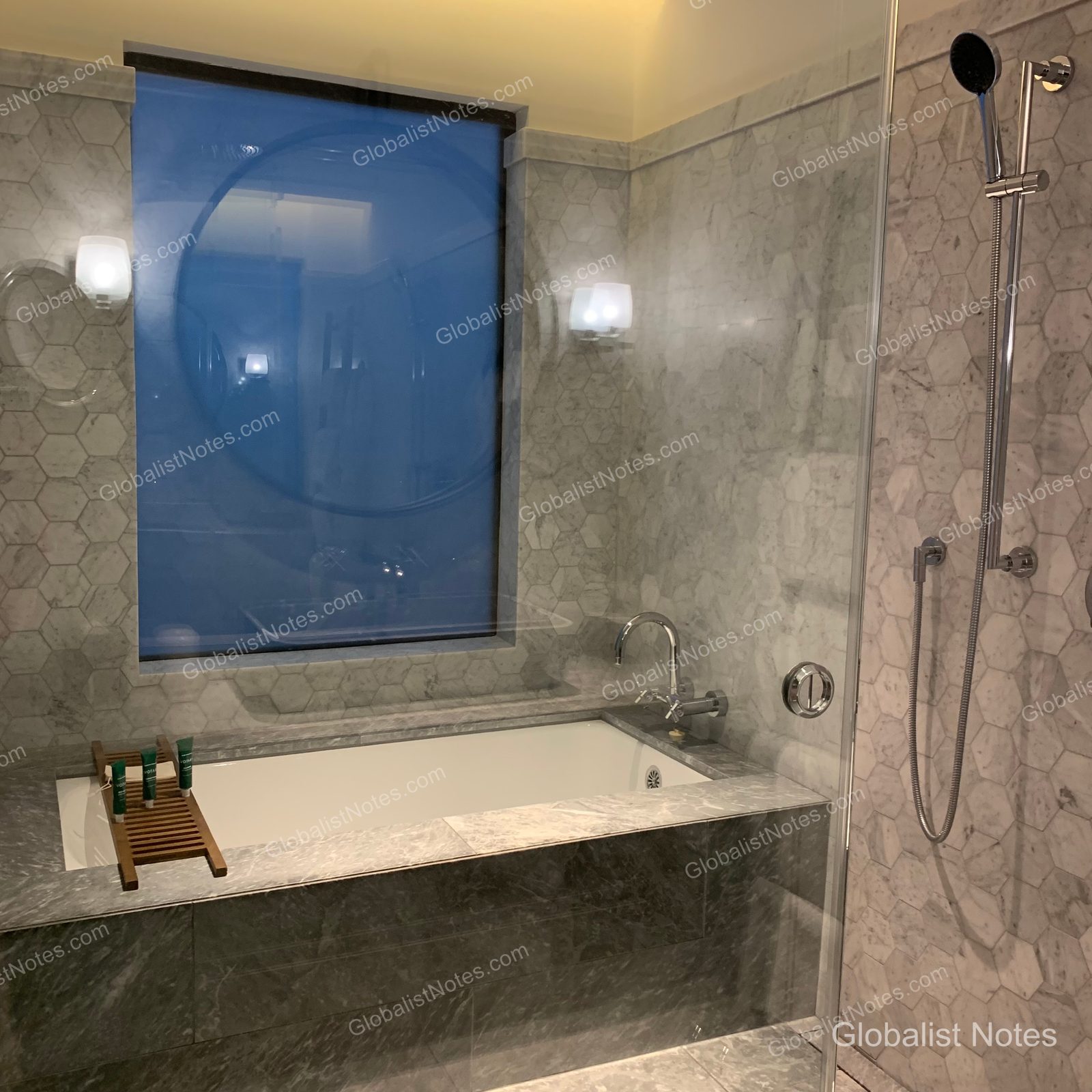 Marble bathroom inside an Alila Napa Valley guest room. A bathtub is positioned in front of a hexagonal tiled wall, with a wooden tray holding amenities placed beside it. To the right is a glass-enclosed shower, and a window with a blue-toned view is located behind the bathtub.