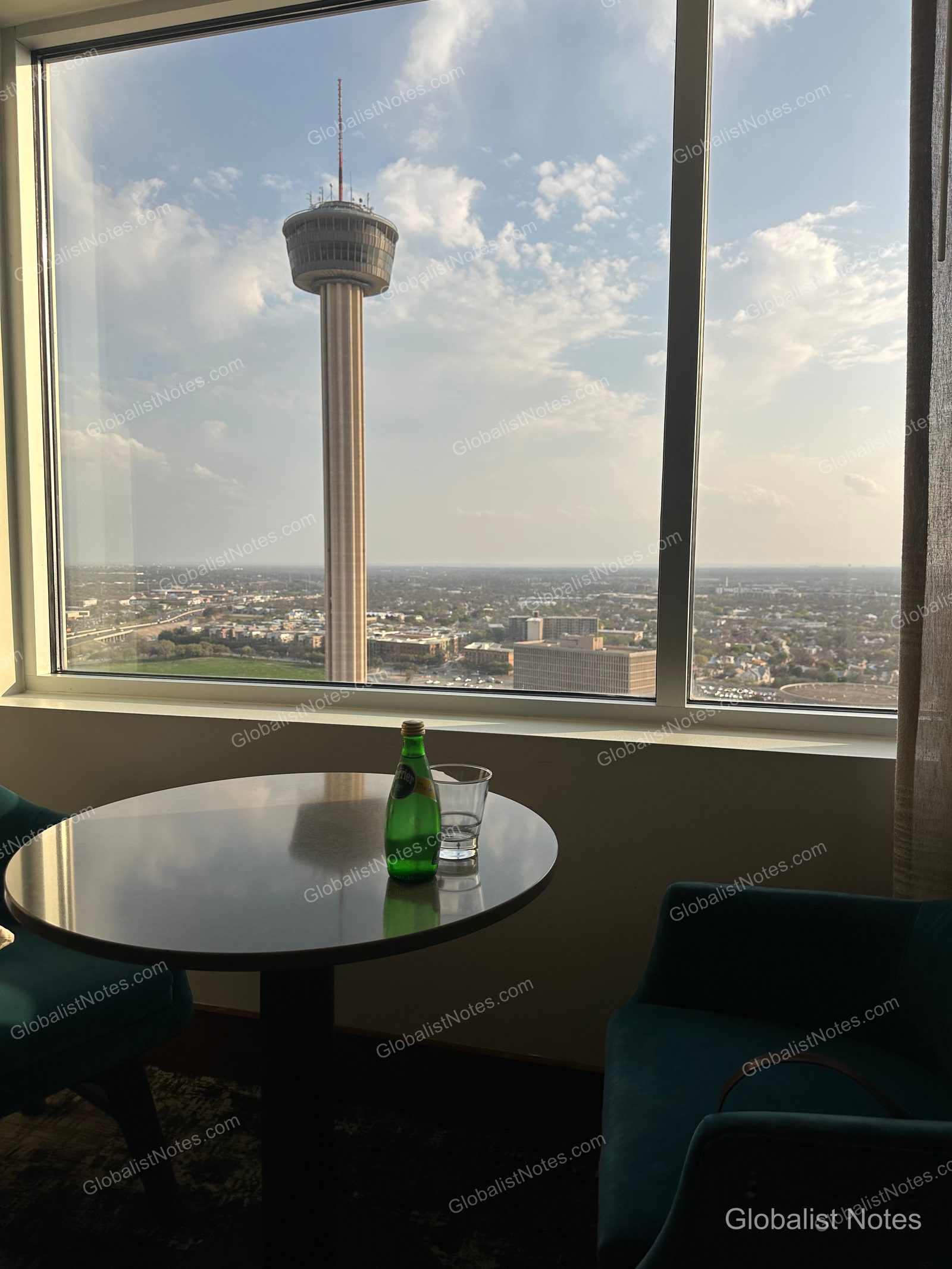 A round table and chair by the guest room window at the Grand Hyatt San Antonio River Walk, with a bottle of Perrier and a glass on the table; the Tower of the Americas is clearly visible against a clear sky outside.