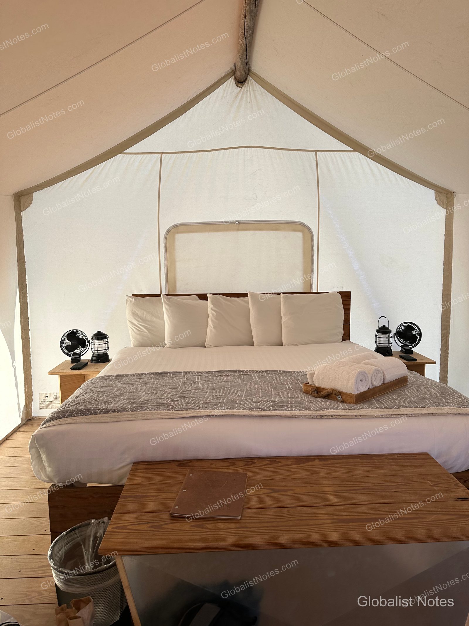 Under Canvas North Yellowstoneのジュニアスイートテント内部。中央にベッドが置かれ、両サイドのテーブルには扇風機とランタンが備えられています。