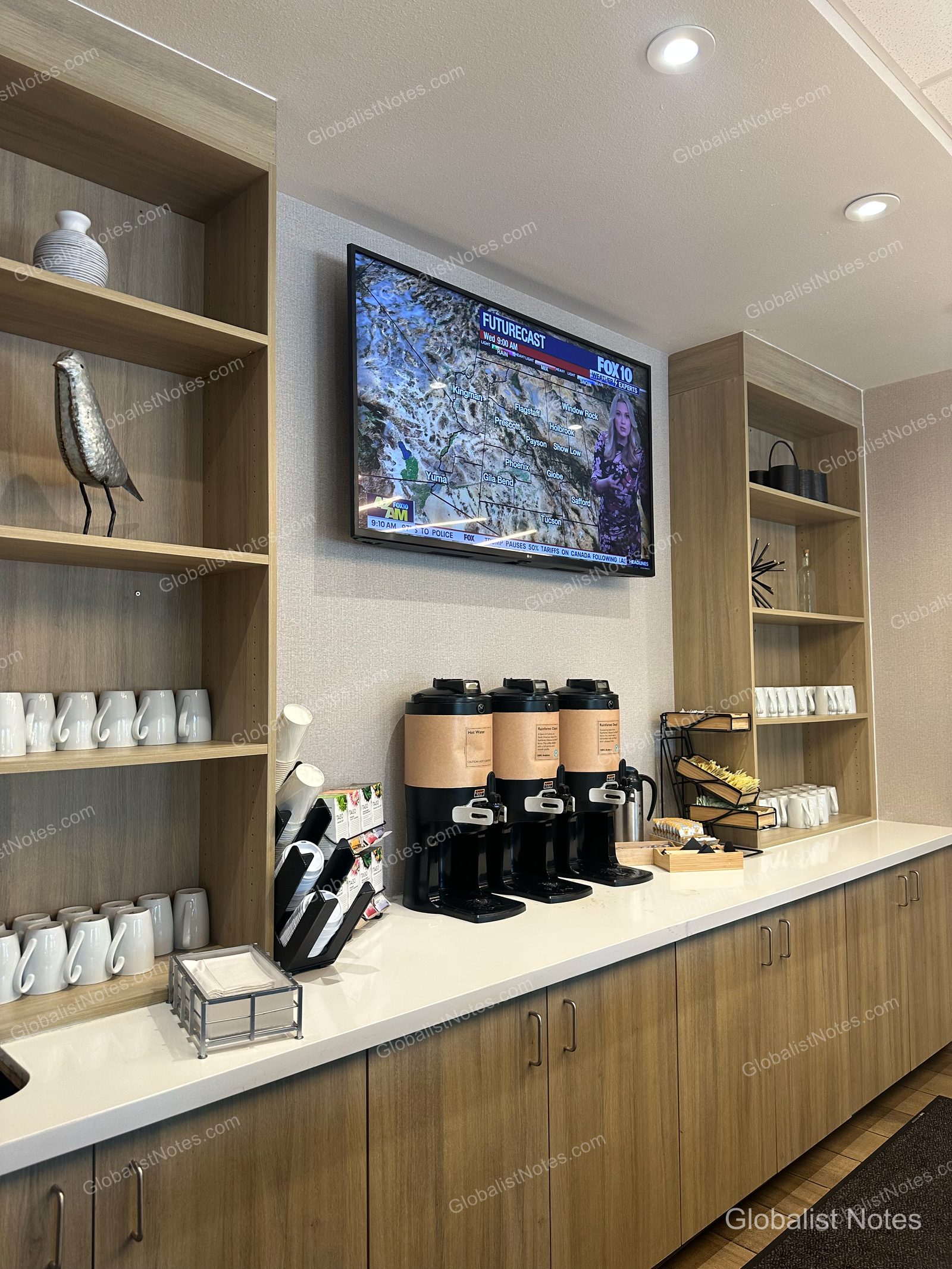 Hyatt Place Flagstaffの朝食エリア。壁掛けテレビの下にコーヒーマシン3台とマグカップ、ティーバッグが整然と並ぶカウンターがあり、両サイドには木製の棚が設置されています。