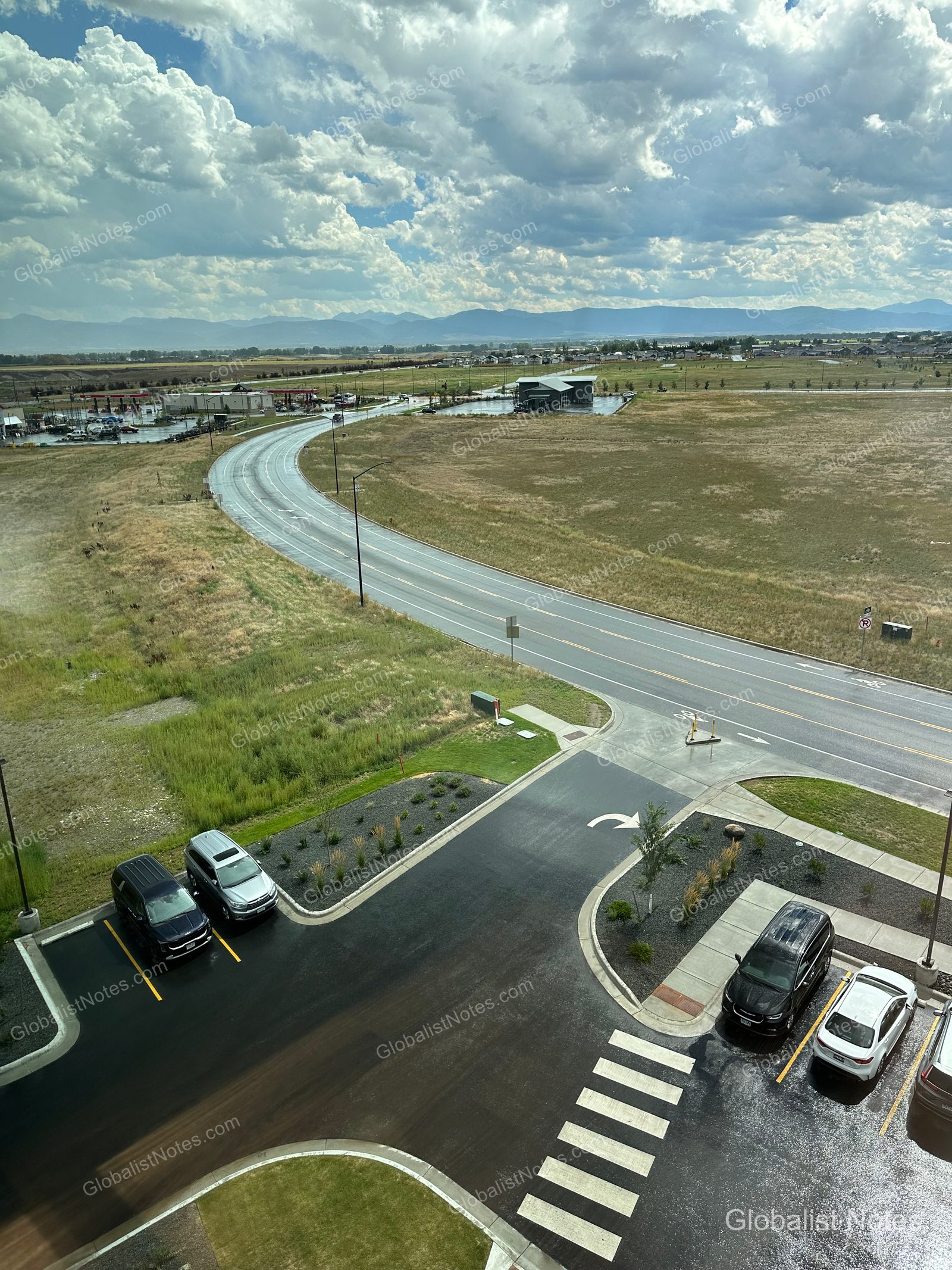 Hyatt Place Bozeman Yellowstone Airportの客室からの眺め。眼下の駐車場と横断歩道の先に、緩やかにカーブする道路と草原、そして雲に覆われた山脈が広がっています。