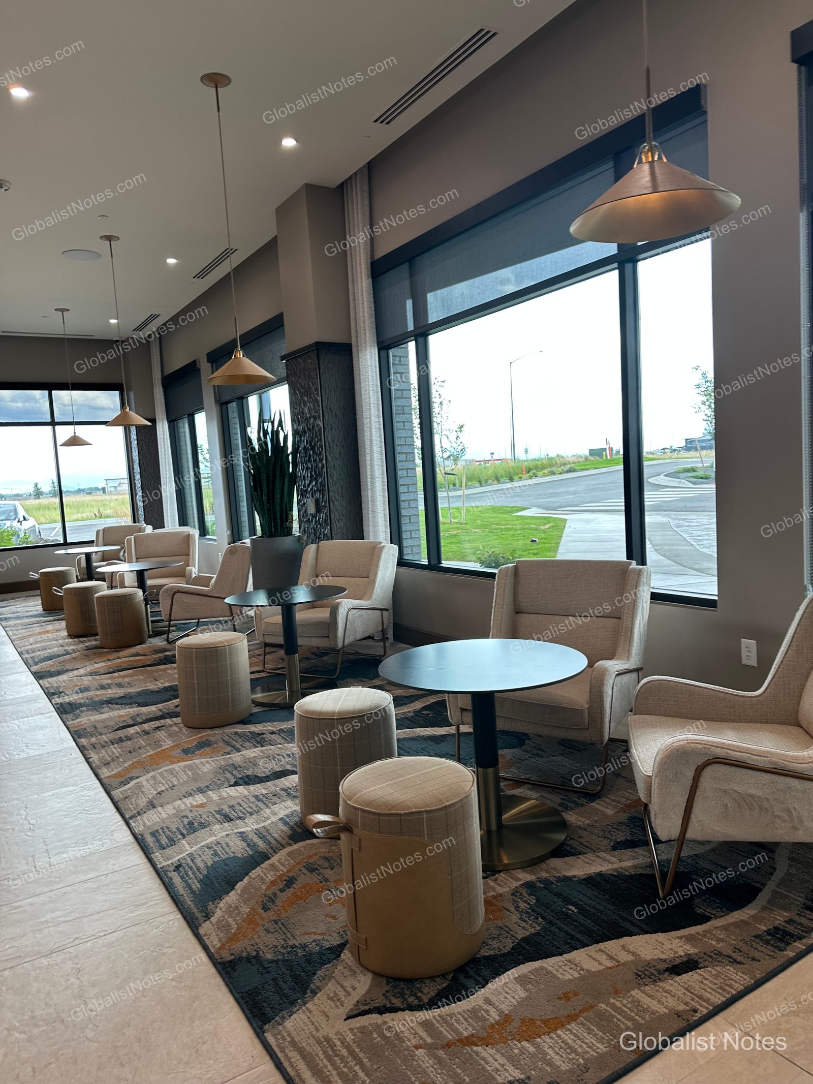 Hyatt Place Bozeman Yellowstone Airportのロビーラウンジ。窓際にベージュのアームチェア、小さな円形テーブル、スツールが並び、床には抽象的なパターンのカーペットが敷かれています。大きな窓からは外の景色が広がります。