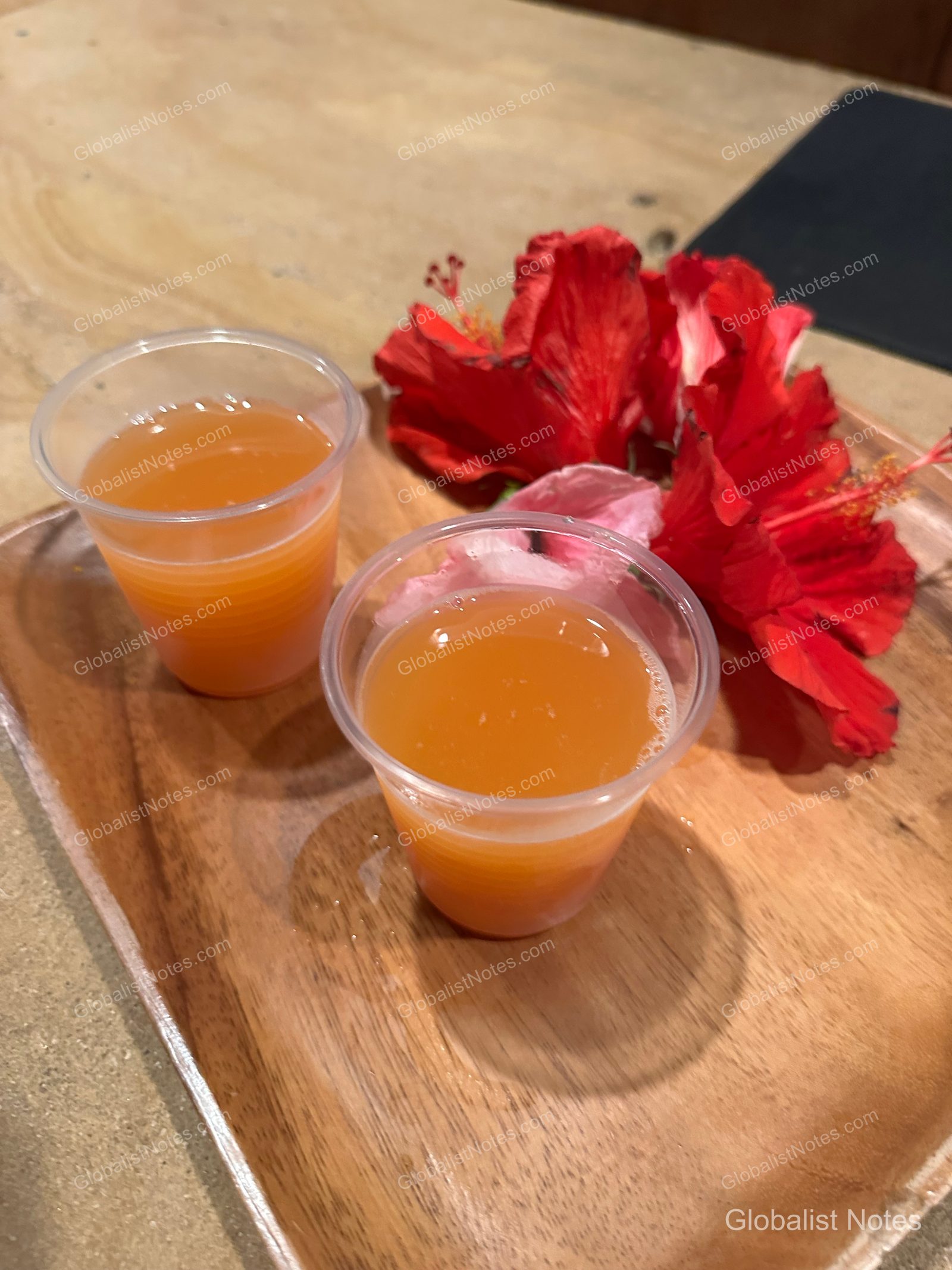Two orange welcome drinks served on a wooden tray at check-in at Hana-Maui Resort, accented with a bright red hibiscus flower.