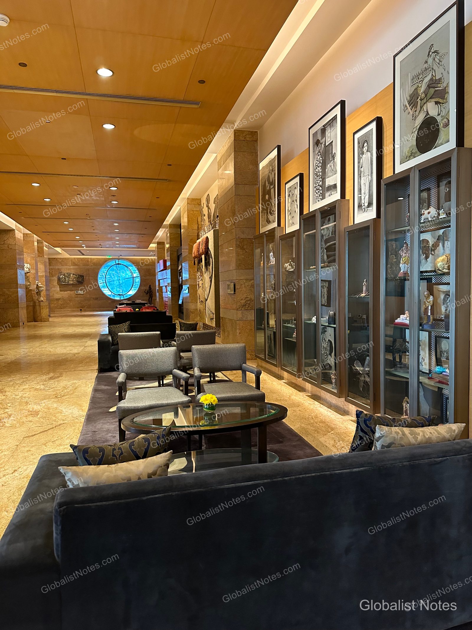 The spacious lobby at Grand Hyatt Mumbai, featuring marble floors, high ceilings, and a seating area with grey armchairs and glass coffee tables. A long wall displays glass cases with art pieces and framed artwork, with a circular window at the end of the corridor.