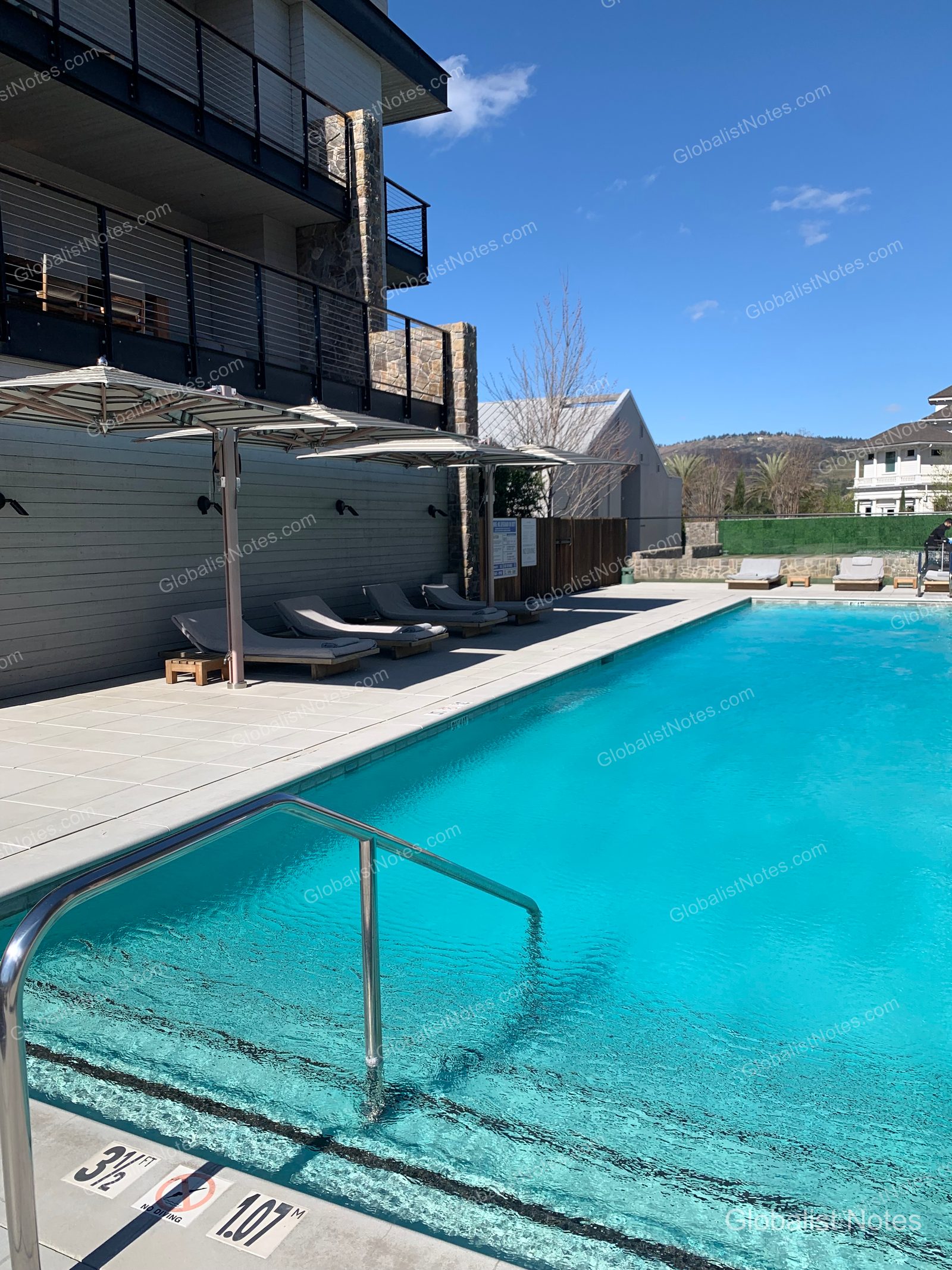 Outdoor pool view at Alila Napa Valley in St. Helena, USA. Sun loungers and umbrellas are lined up beside the bright blue pool, with modern hotel guest room buildings in the background.