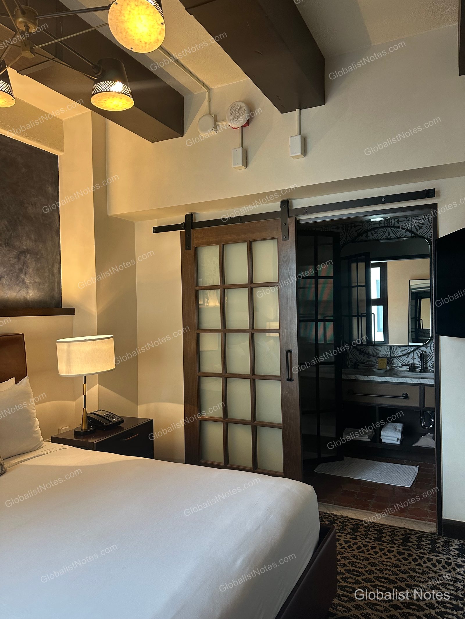Interior of a Junior Suite at Hotel Figueroa, Unbound Collection by Hyatt in Los Angeles. A bedside table and lamp are visible, with a lattice-style sliding door leading to the bathroom vanity and mirror.