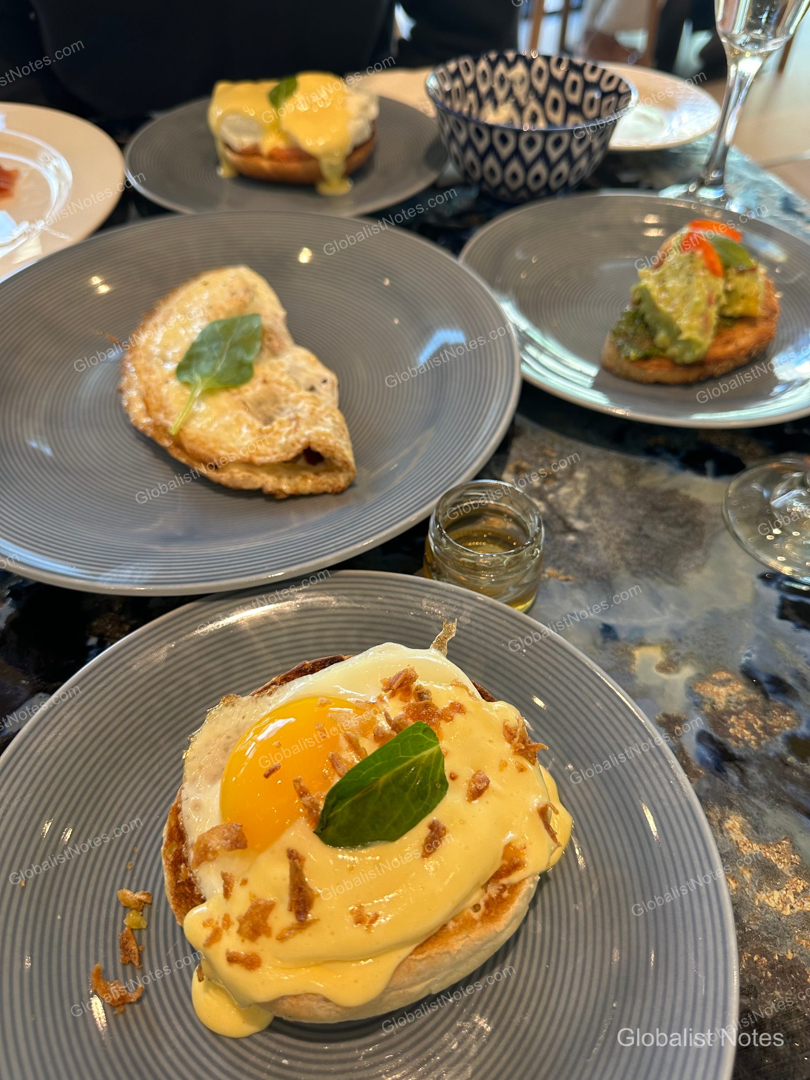 Breakfast spread at Hyatt Regency Malta featuring Eggs Benedict, an omelet, and avocado toast on grey plates, served with a small jar of honey.