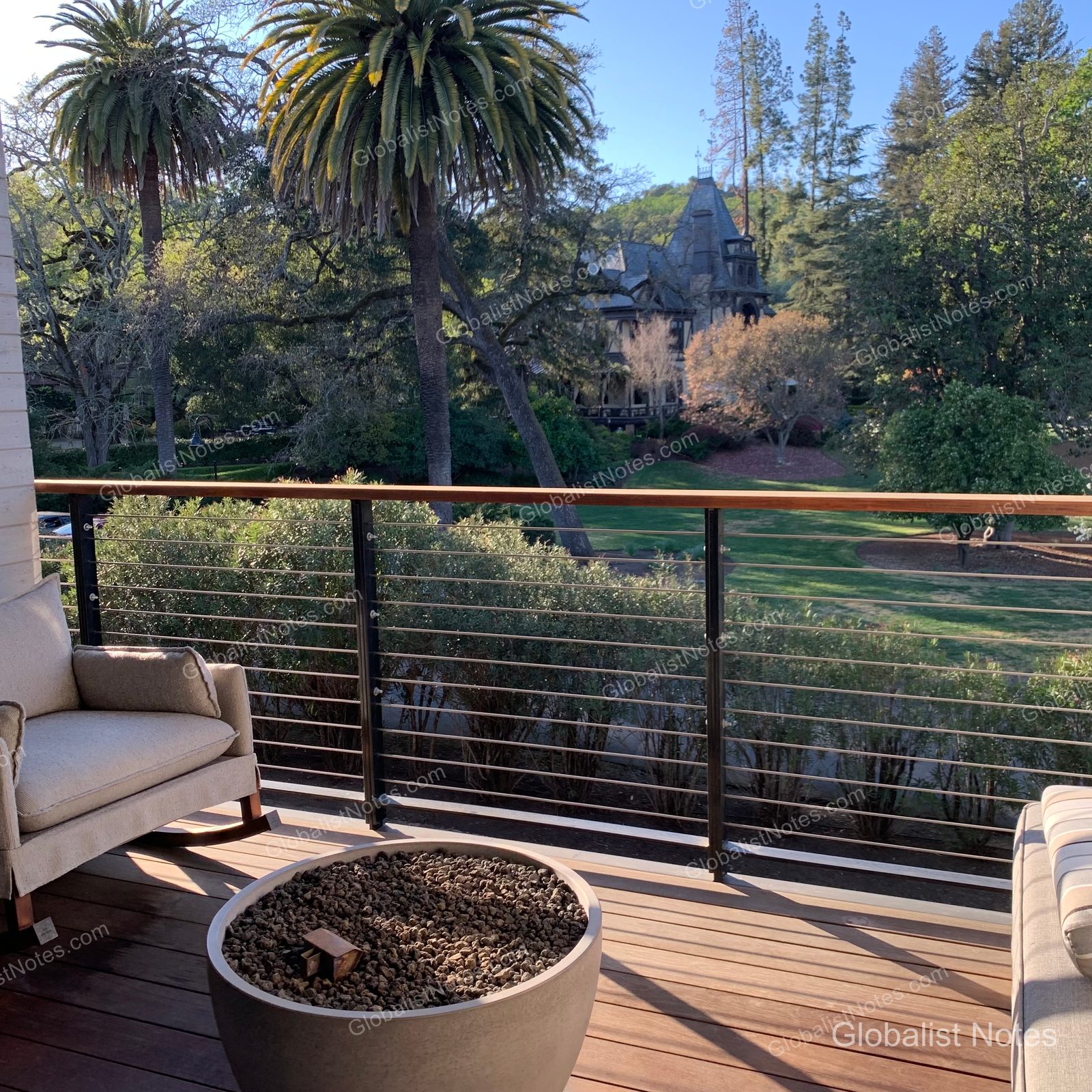 Terrace of a 2 Queen Bed Vineyard View Balcony room at Alila Napa Valley. The wooden deck features a circular outdoor fire pit and beige cushioned chairs, with a view over the railing of green vineyards, lush trees, and historic buildings in the distance.
