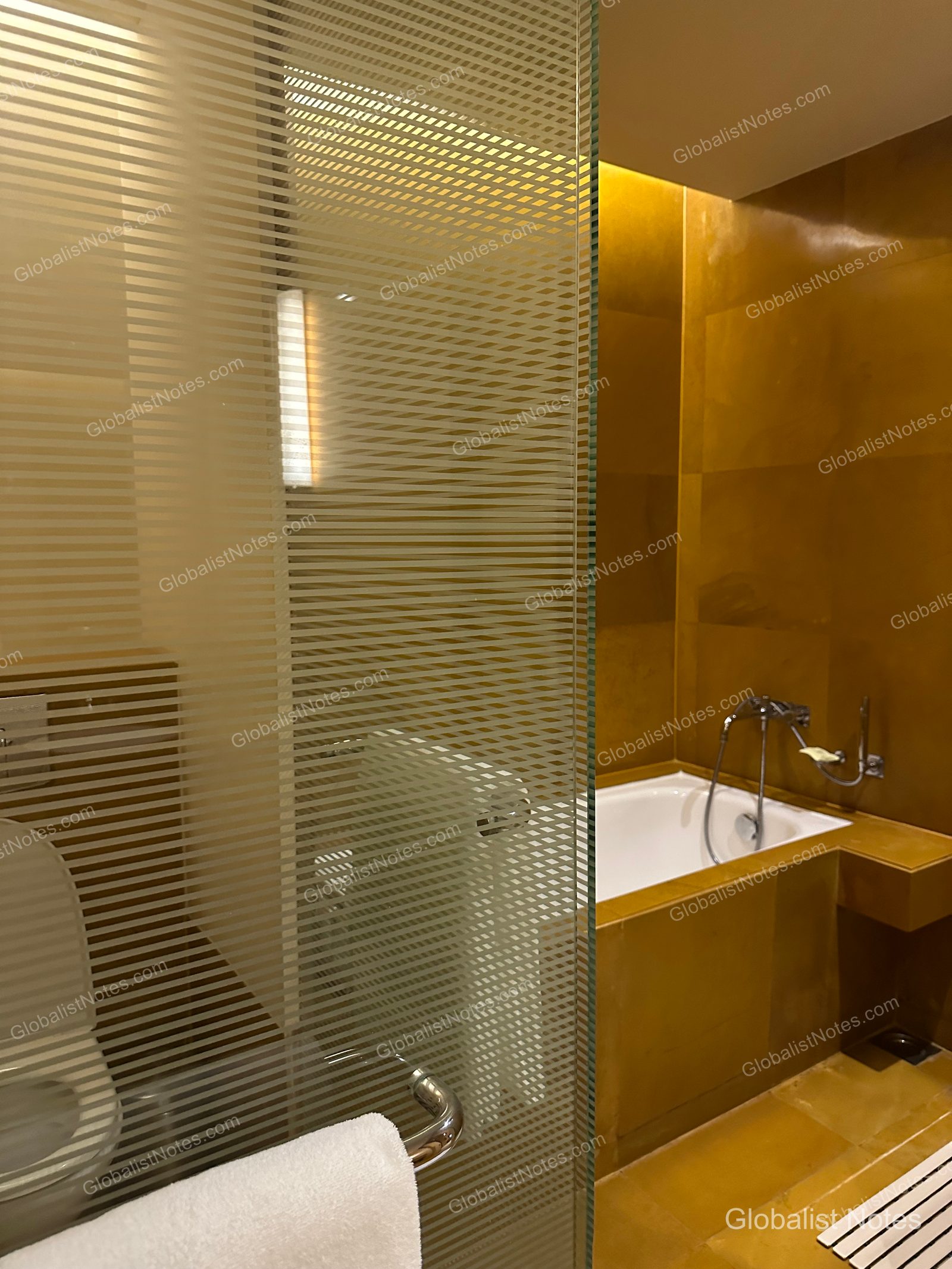 Bathroom of the Veranda Suite King at Grand Hyatt Mumbai Hotel and Residences. A translucent, striped glass partition separates the space, revealing a bathtub and shower area finished with gold-toned tiles and white towels.