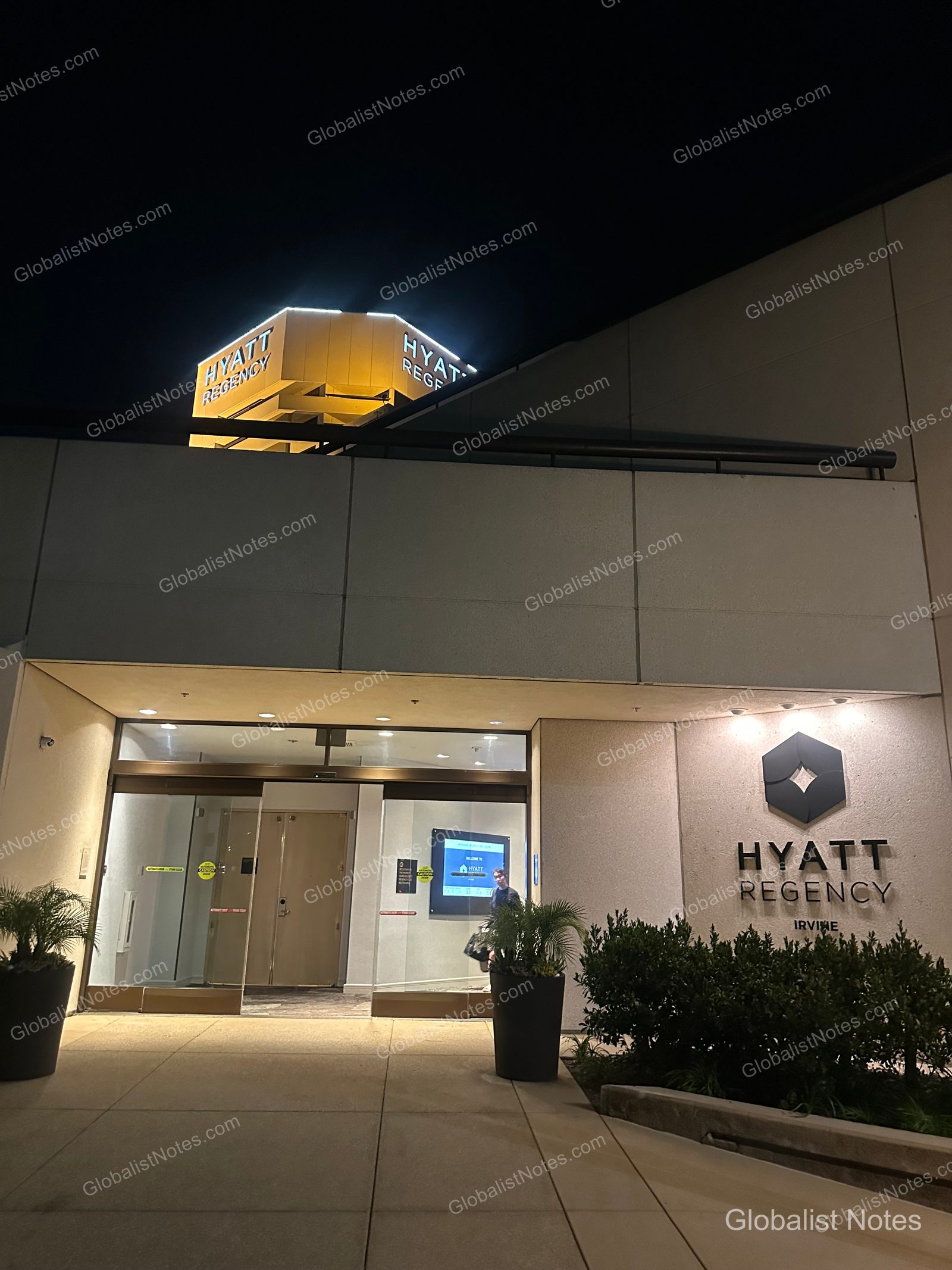 夜間點燈後的 Hyatt Regency Irvine 入口。建築上方有明亮的 Hyatt Regency 標誌，入口旁牆面亦設有飯店名稱與 Logo。門口擺放盆栽，透過自動門可隱約看見大廳內部。
