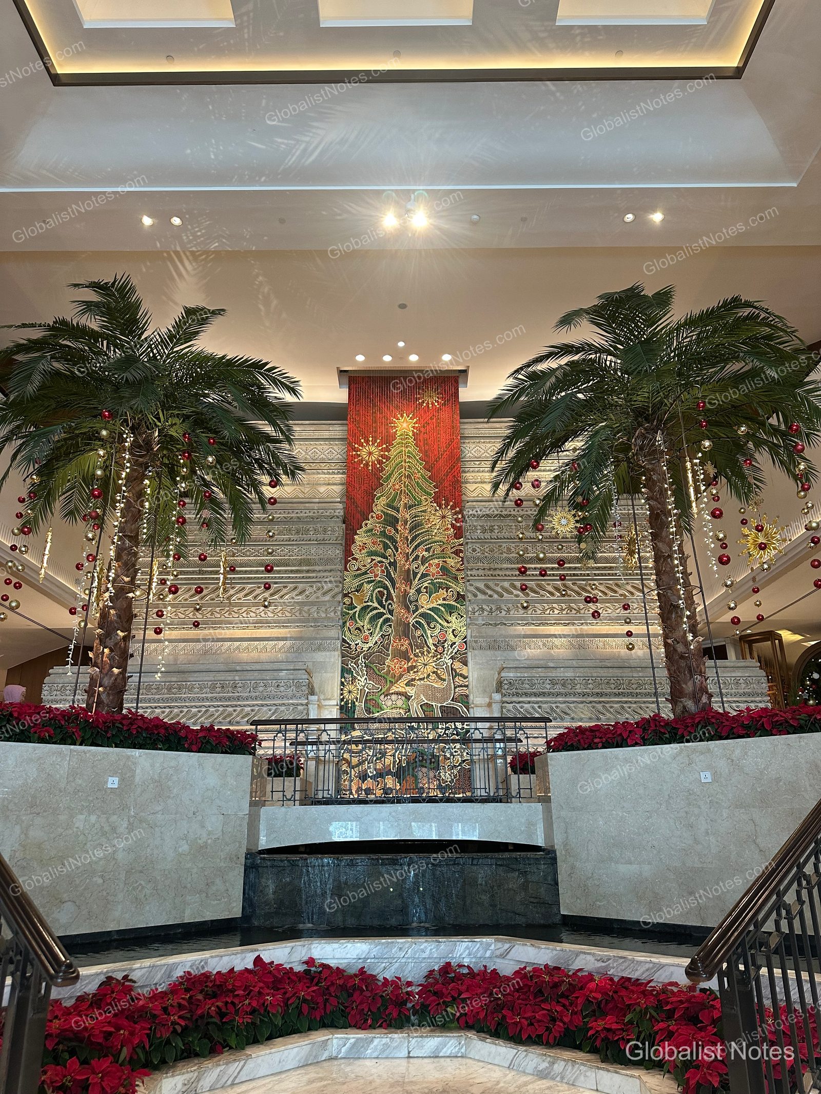 A large Christmas tree stands in the center of the Grand Hyatt Jakarta lobby, flanked symmetrically by two illuminated palm trees against a marble wall, with red poinsettias placed at the base.