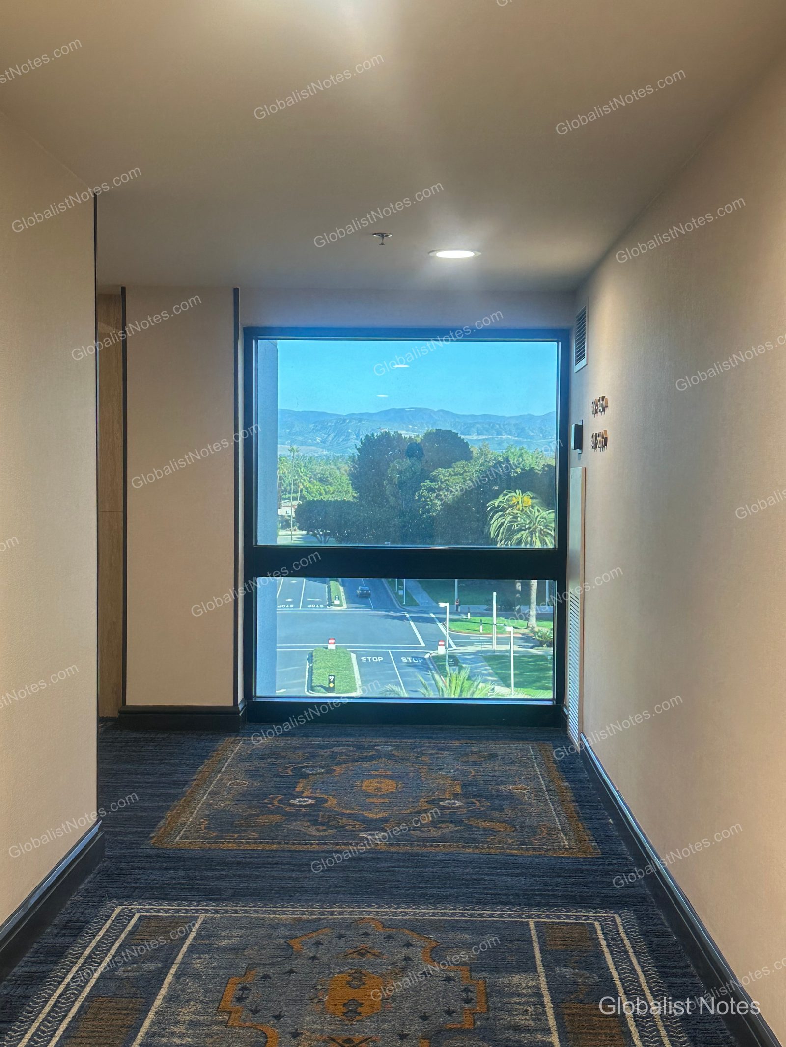 Hyatt Regency Irvine 飯店走廊盡頭的大面窗景，可眺望爾灣街景與遠方山巒。地面鋪設花紋地毯，窗邊牆面設有客房編號指示牌。
