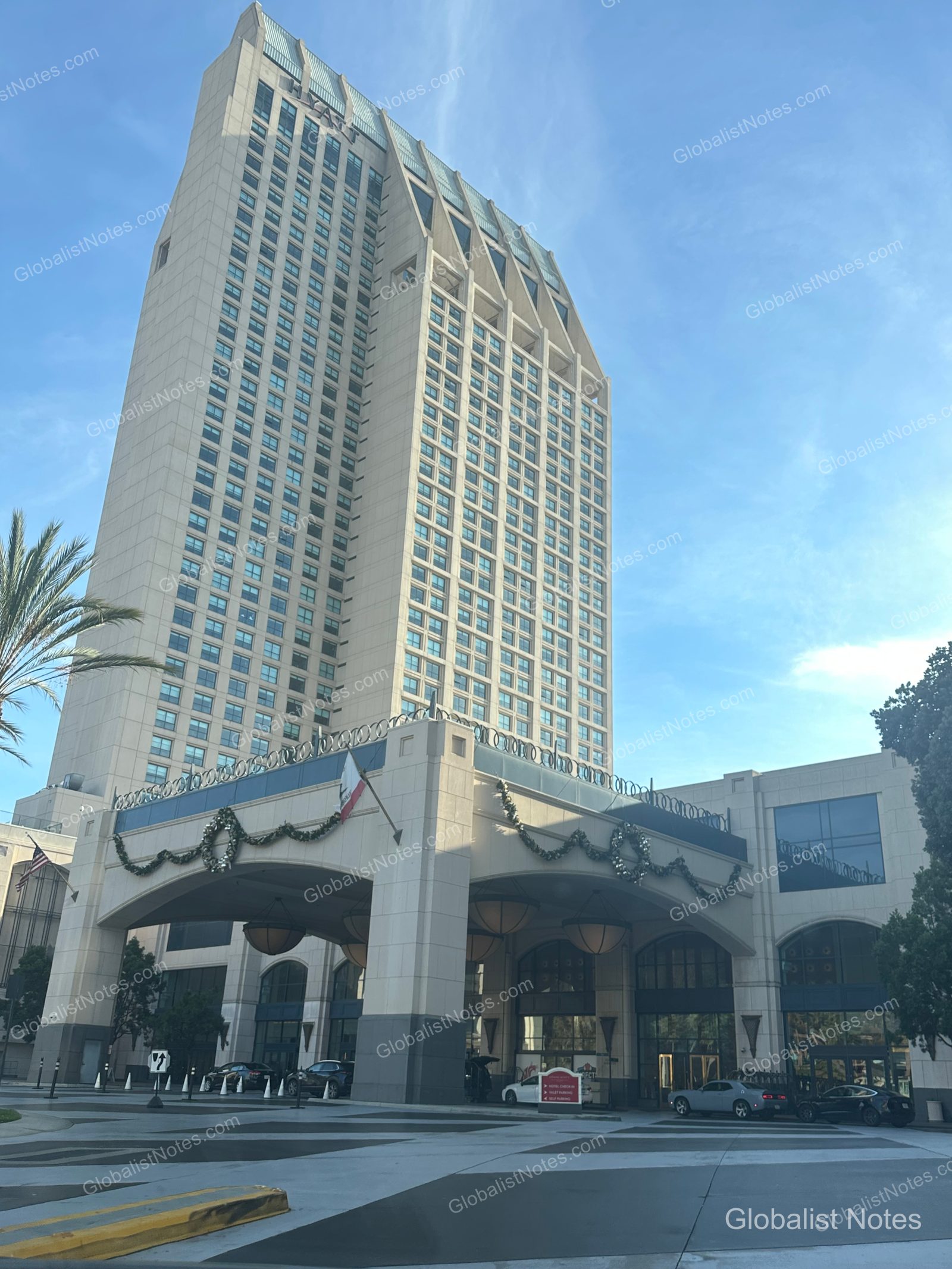 Manchester Grand Hyatt San Diego 外觀，入口處裝飾著聖誕節慶裝飾，高聳的建築在晴空下相當醒目，門口有車輛停靠，一側種植著棕櫚樹。