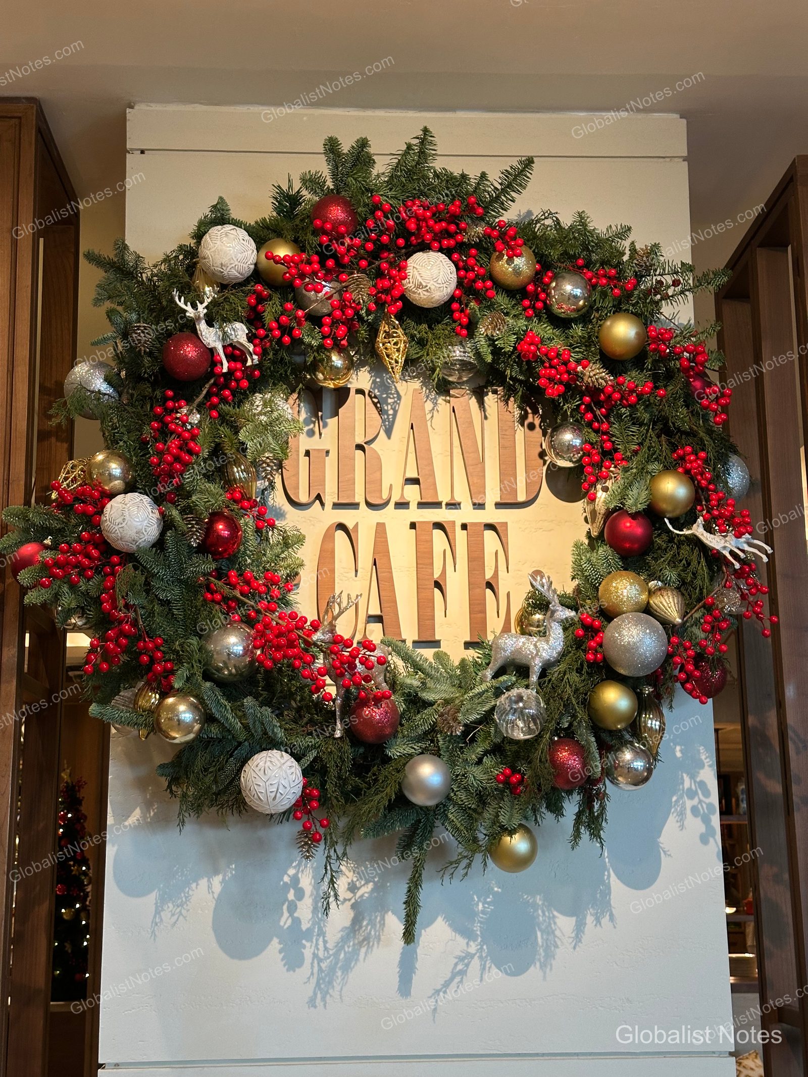 A large Christmas wreath hanging at the entrance of Grand Café at Grand Hyatt Jakarta, decorated with green pine branches, red berries, gold and silver ornaments, and deer figures.