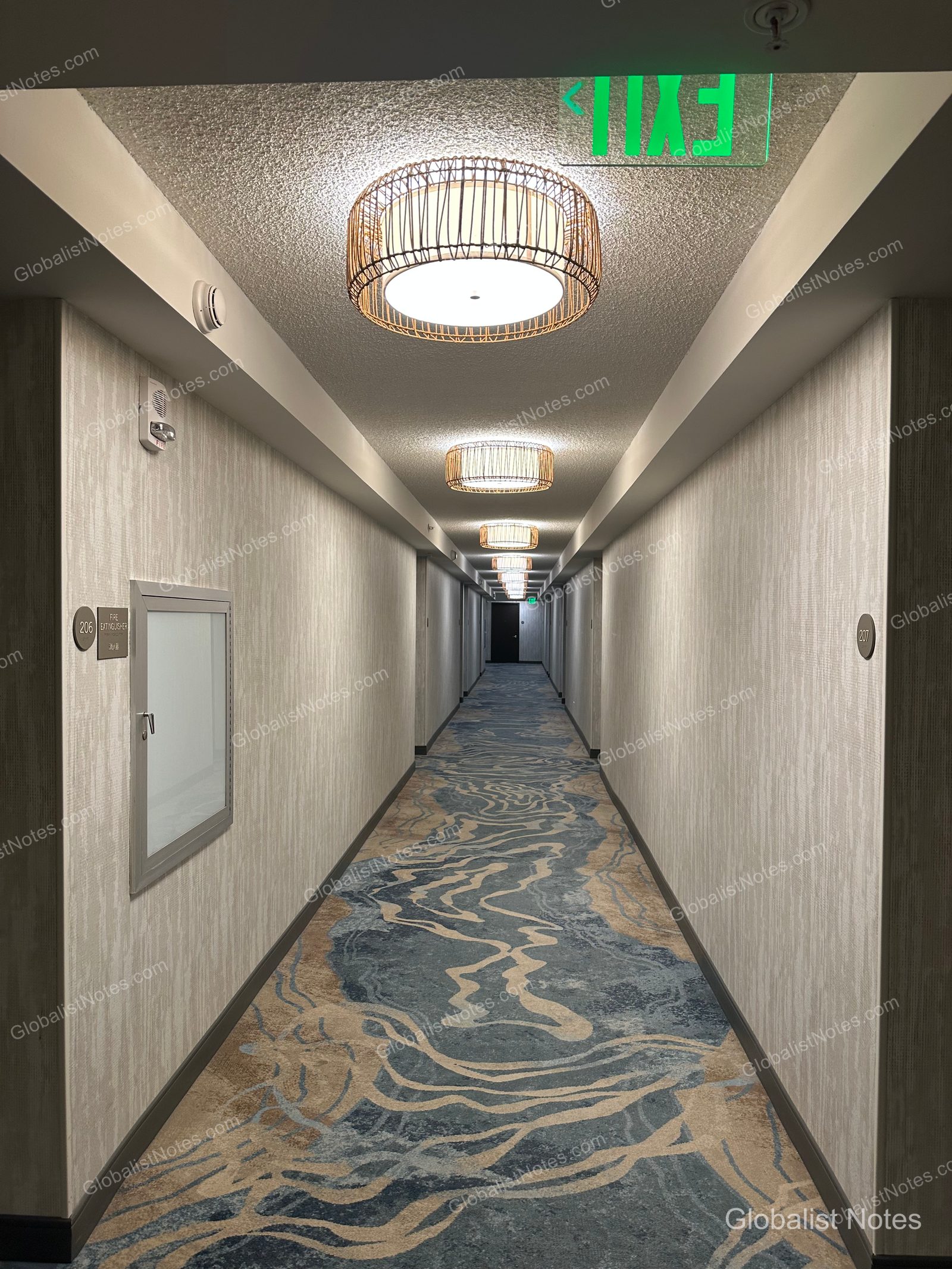 A hallway at Hyatt Place Waikiki Beach in Hawaii, featuring blue and beige wave-patterned carpeting, a row of circular rattan ceiling lights, and room signage on the walls.