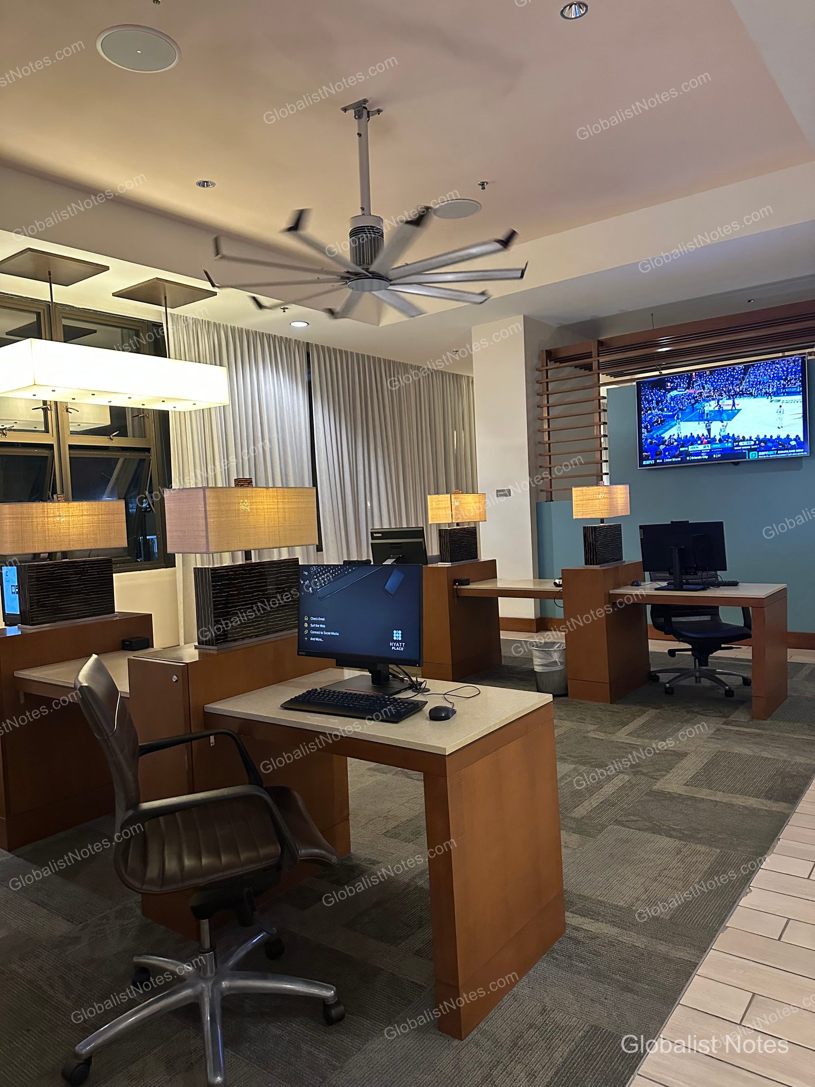 Business center at Hyatt Place Waikiki Beach in Honolulu, Hawaii. Individual desks with computers and chairs are arranged in the space, with a large ceiling fan overhead and a TV playing sports mounted on the wall.
