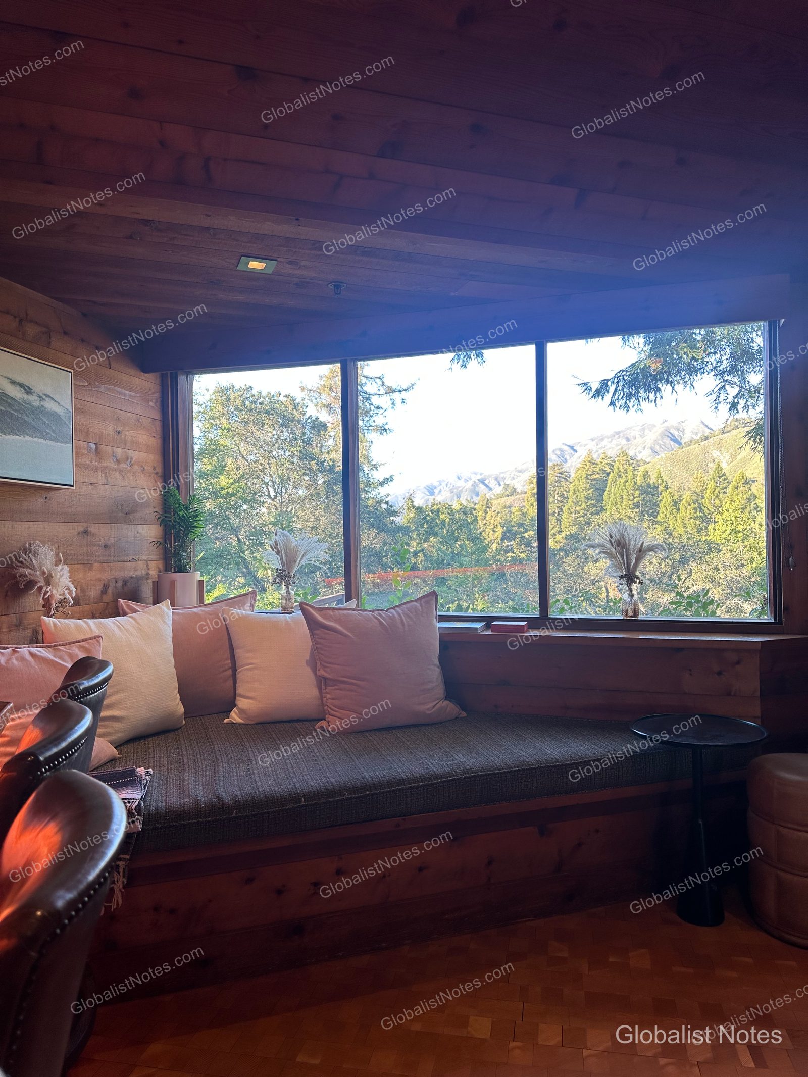A long bench-style sofa with soft cushions placed by the window in an Alila Ventana Big Sur guest room. Outside the window, the lush forests and mountain ranges of Big Sur are visible, and the interior features wood-paneled walls.