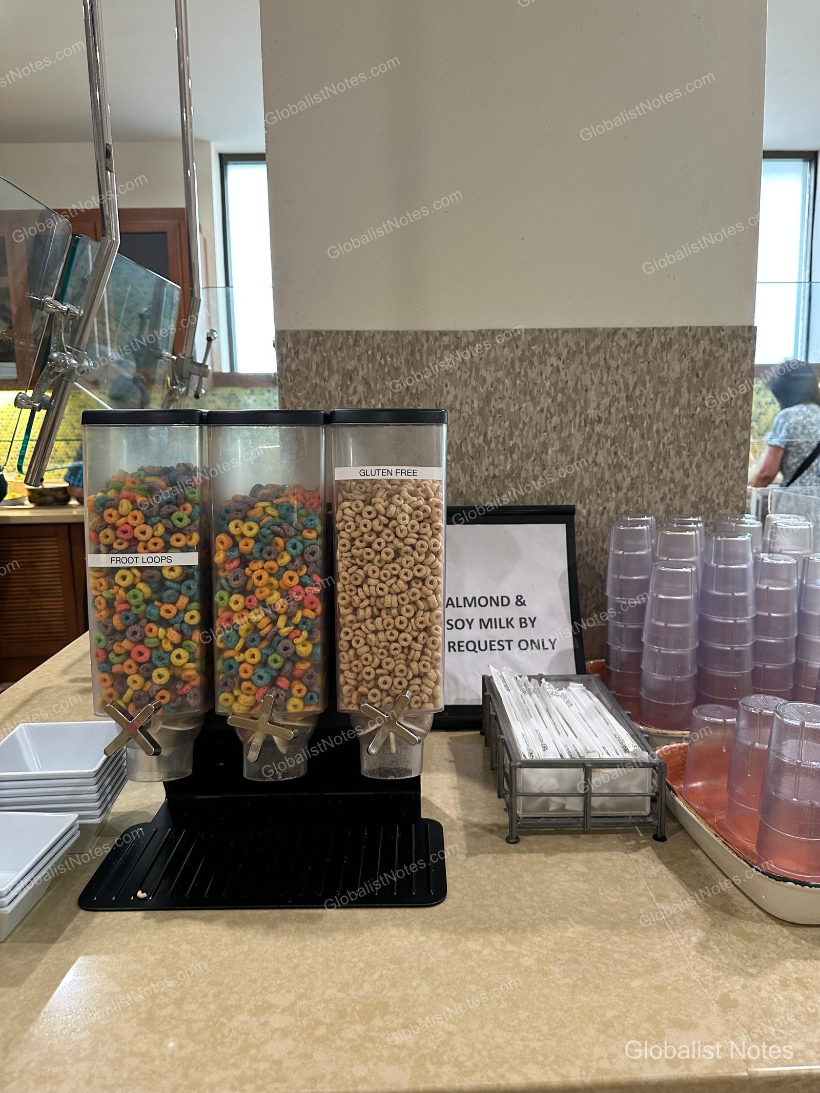 Three cereal dispensers at the Hyatt Place Waikiki Beach breakfast area. From left, two contain Froot Loops and one contains gluten-free cereal. Next to them is a sign stating that almond and soy milk are available upon request, along with plastic cups and straws.