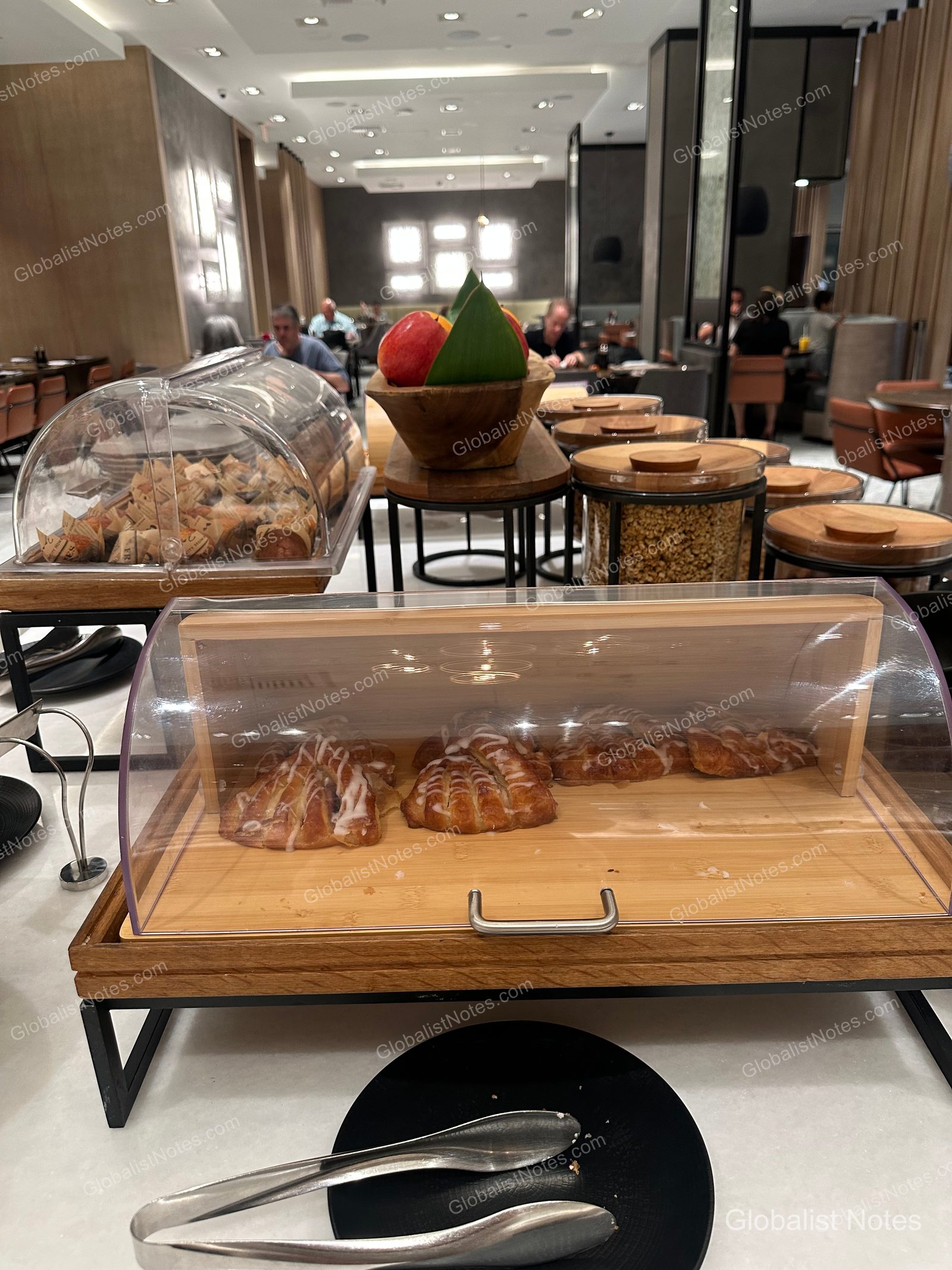 Hyatt Regency Newport Beach 早餐自助餐區。透明蓋木製托盤內擺放著淋上糖霜的糕點，後方則有裝滿蘋果的木碗與麥片罐。