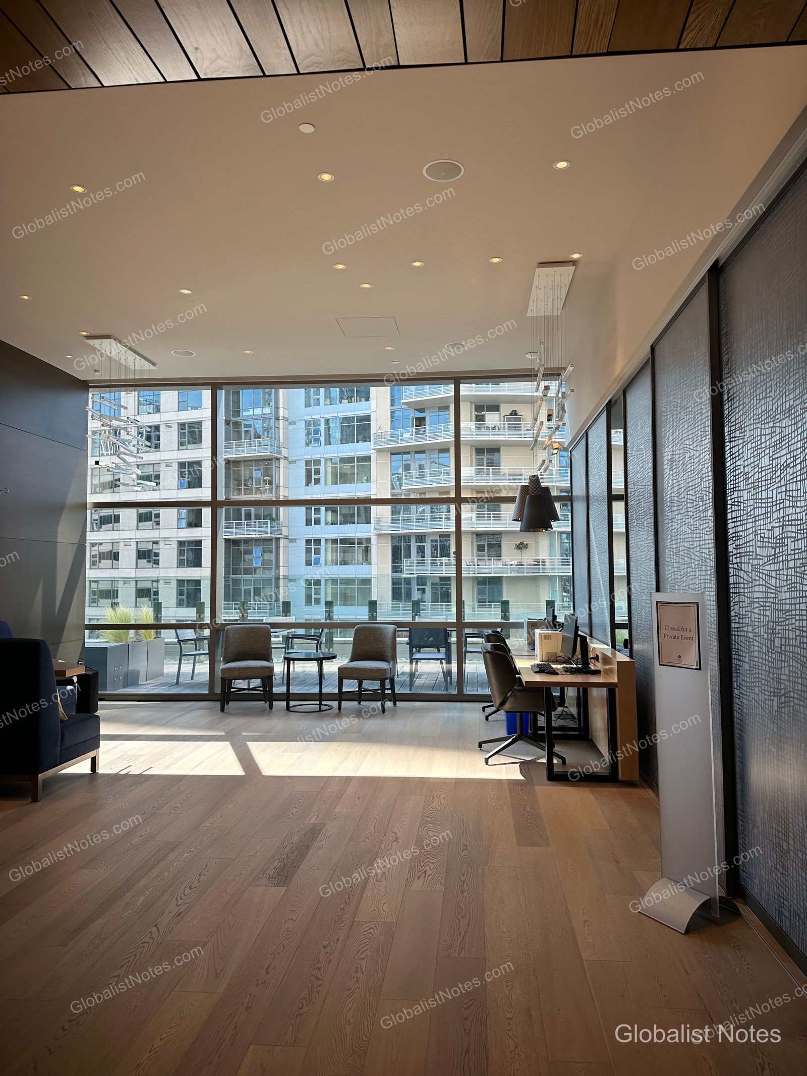 Lounge area at Hyatt Regency Seattle featuring floor-to-ceiling windows with views of nearby buildings, light wood flooring, and arranged chairs and desks. The ceiling includes recessed and pendant lighting, with an information sign positioned on the right.