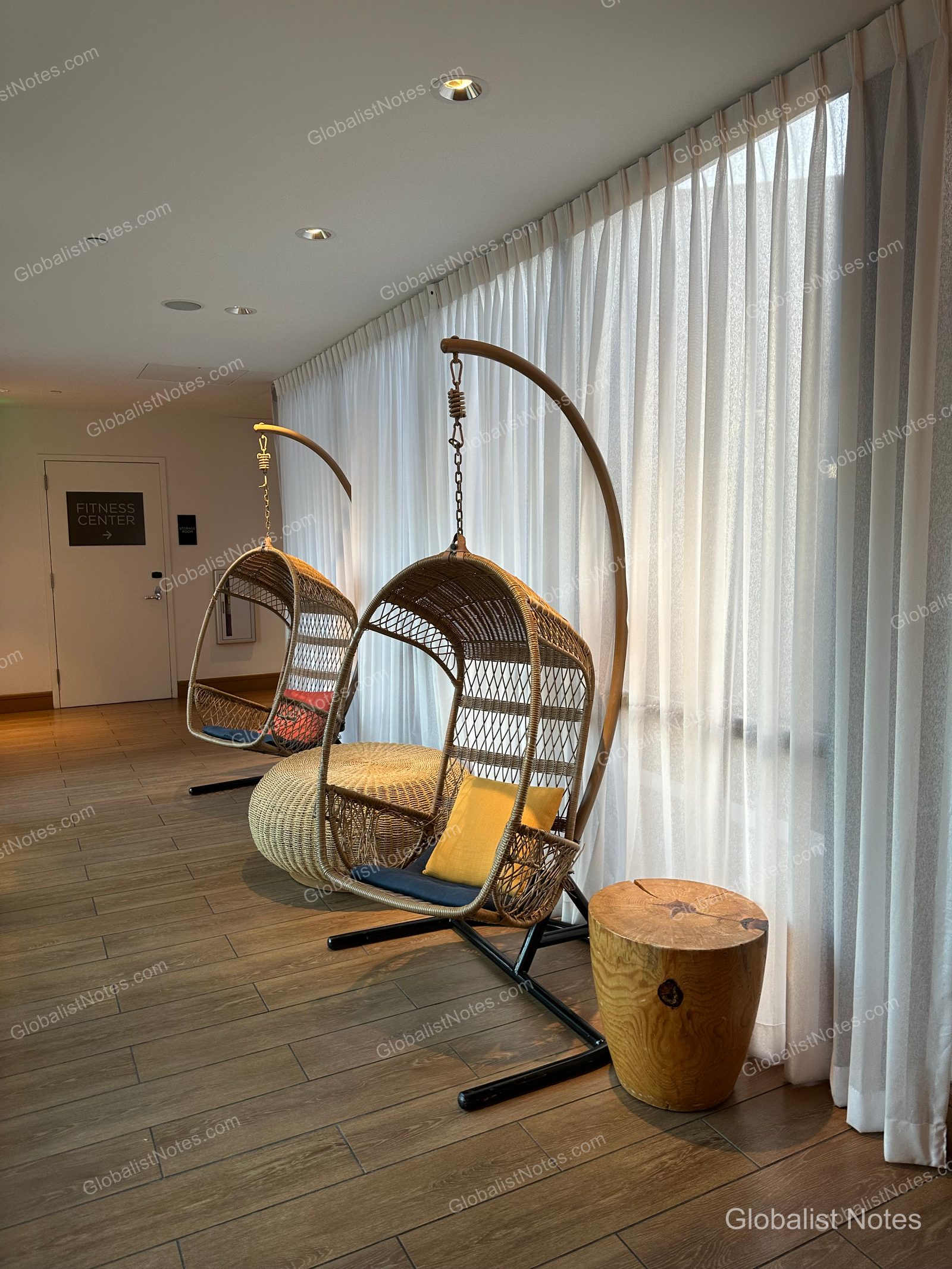 Two rattan rocking chairs placed side-by-side in the hallway next to the fitness center entrance at Hyatt Centric Waikiki Beach. A tree stump-style side table sits between the chairs, set against a backdrop of floor-to-ceiling windows draped with white curtains.