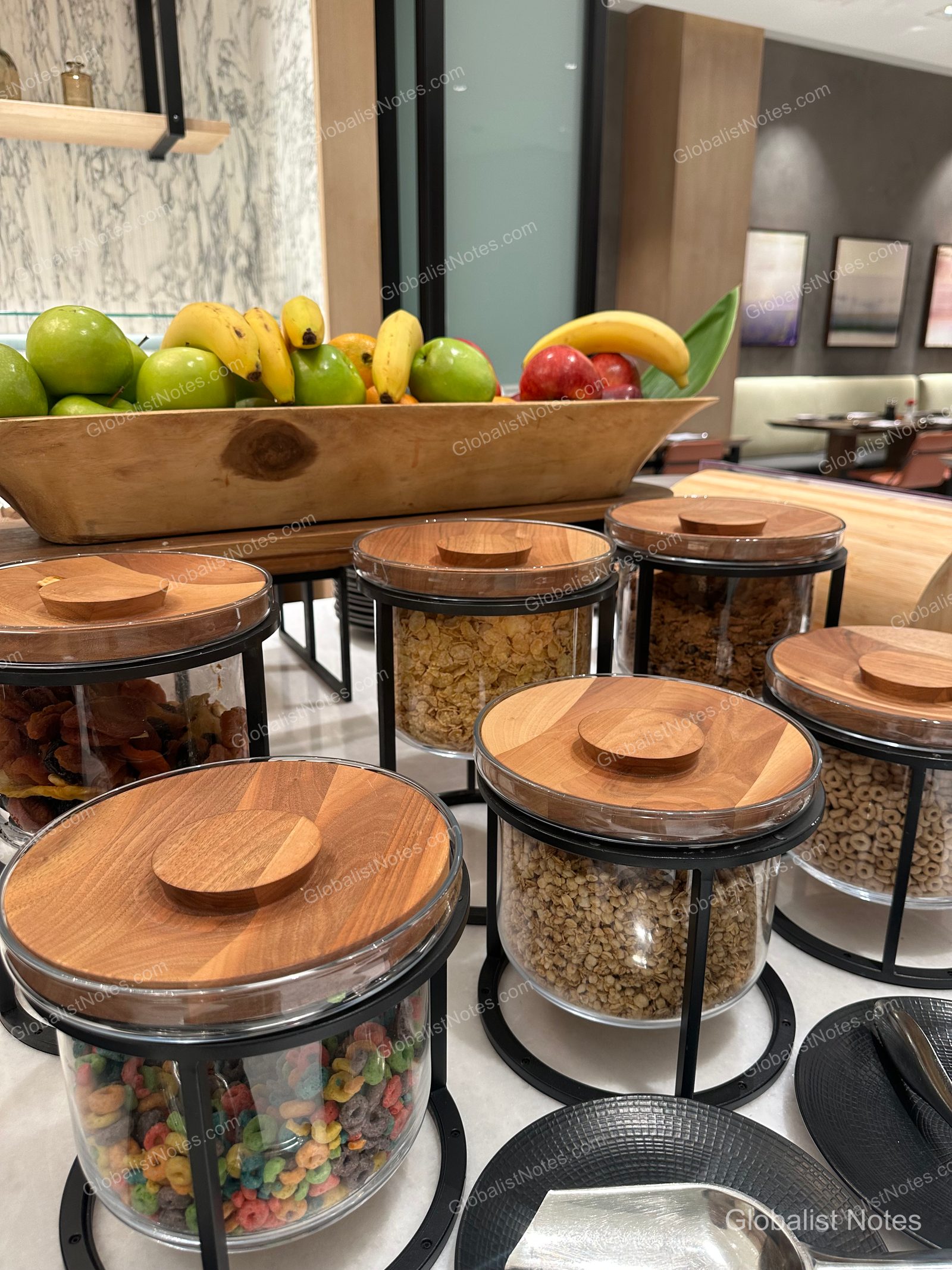 Hyatt Regency Newport Beach West 早餐自助餐的麥片分配器與水果籃。多個木蓋透明玻璃罐裝有不同種類麥片，後方長木碗則盛裝著香蕉與蘋果等新鮮水果。