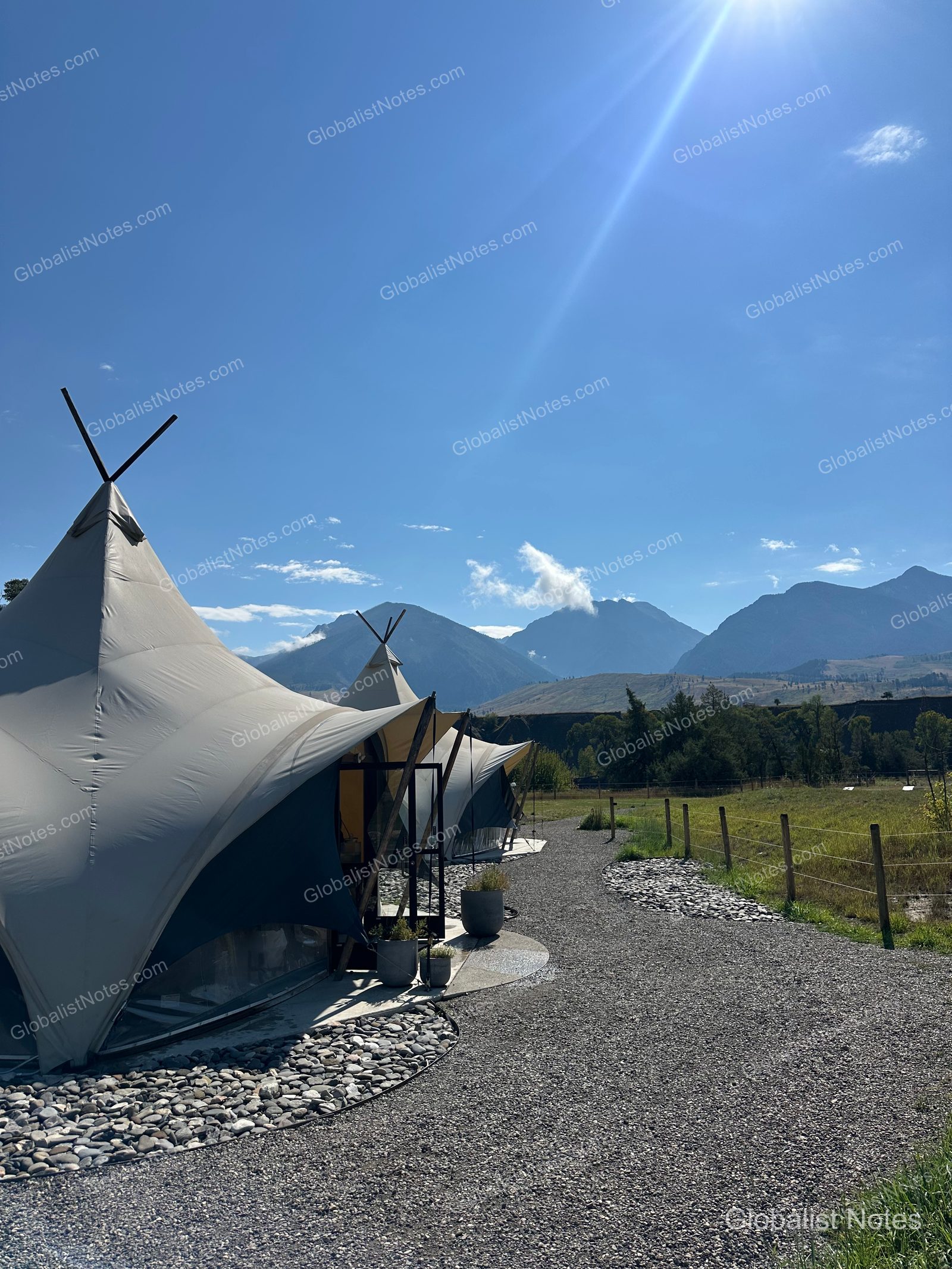 Under Canvas North Yellowstoneのテント宿泊エリア。砂利道の両側に円錐形のテントが並び、背後には晴天の下、雄大な山脈がそびえ立つ。