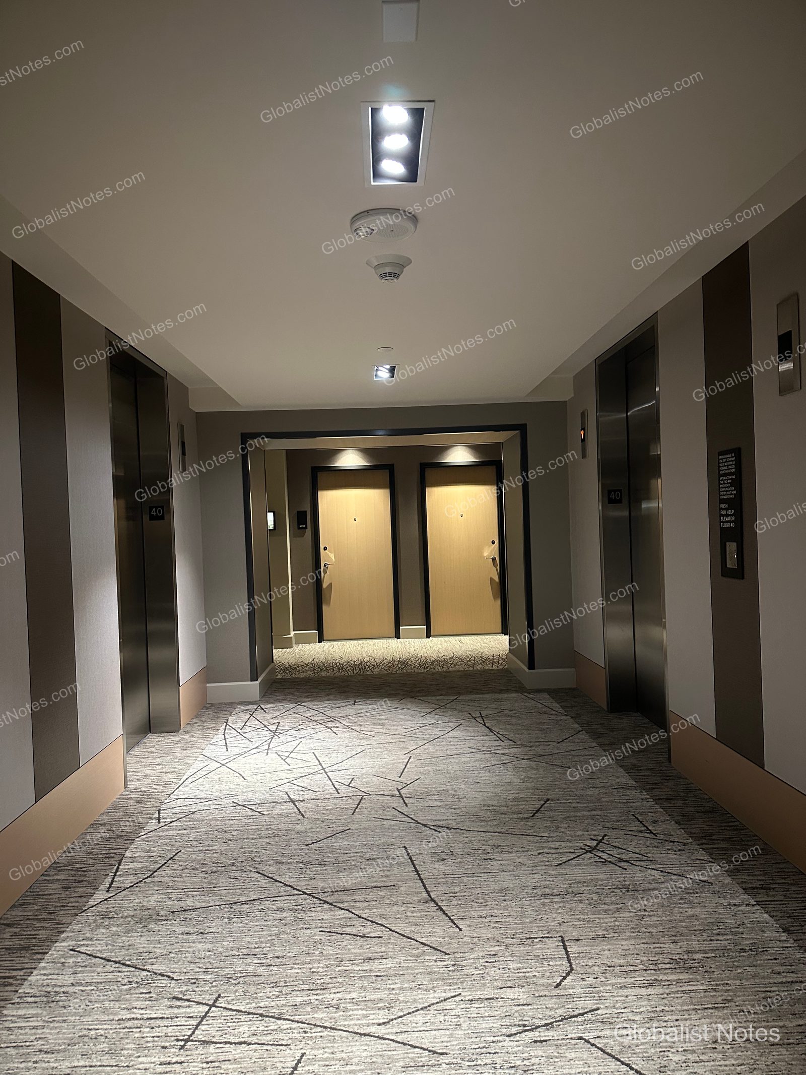 Guest room floor hallway at Hyatt Regency Seattle. Silver elevators are located on both sides, with two guest room doors visible at the end of the central corridor. The floor is covered in a gray carpet with a black linear pattern, and recessed lighting in the ceiling creates a calm atmosphere.