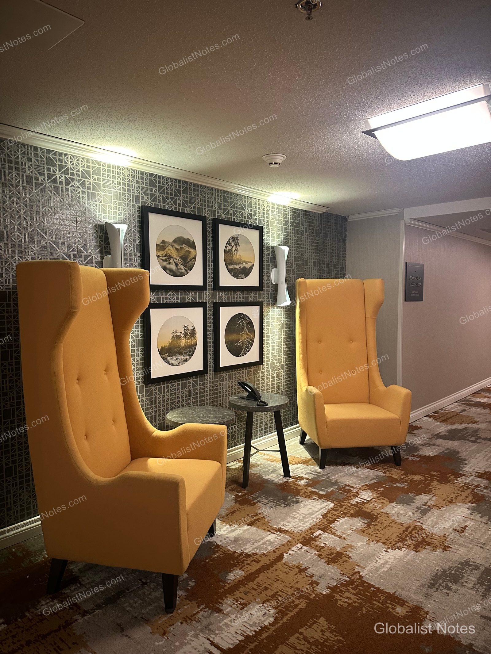 Geometric patterned wallpaper and four circular framed artworks line the hallway of the Hyatt Regency Vancouver, with two yellow high-back armchairs and a small round table placed below.