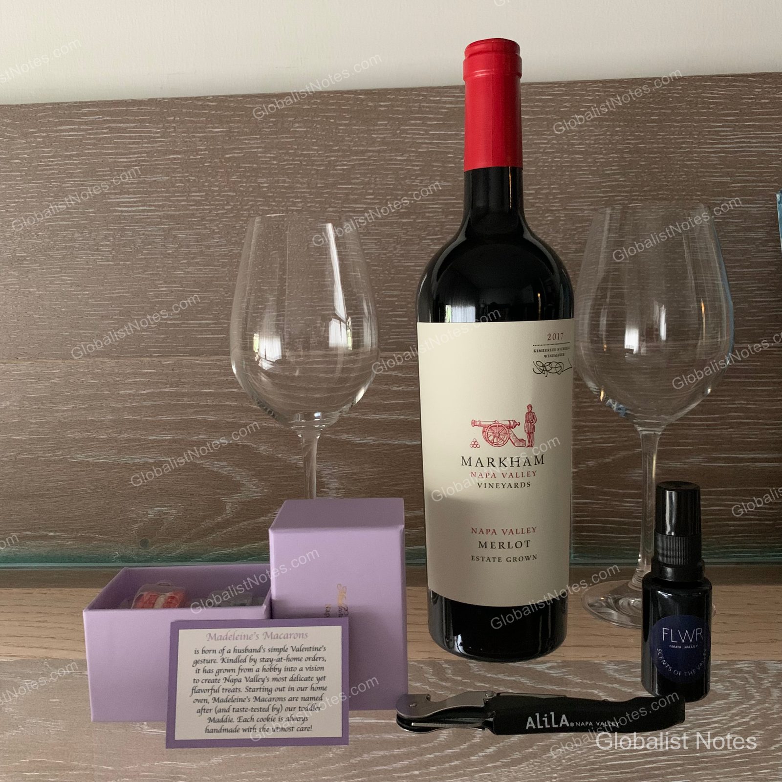 A bottle of Markham Merlot, two wine glasses, a purple box of madeleines and macarons, an Alila-branded wine opener, and a small perfume bottle neatly arranged on the wooden table inside an Alila Napa Valley guest room.