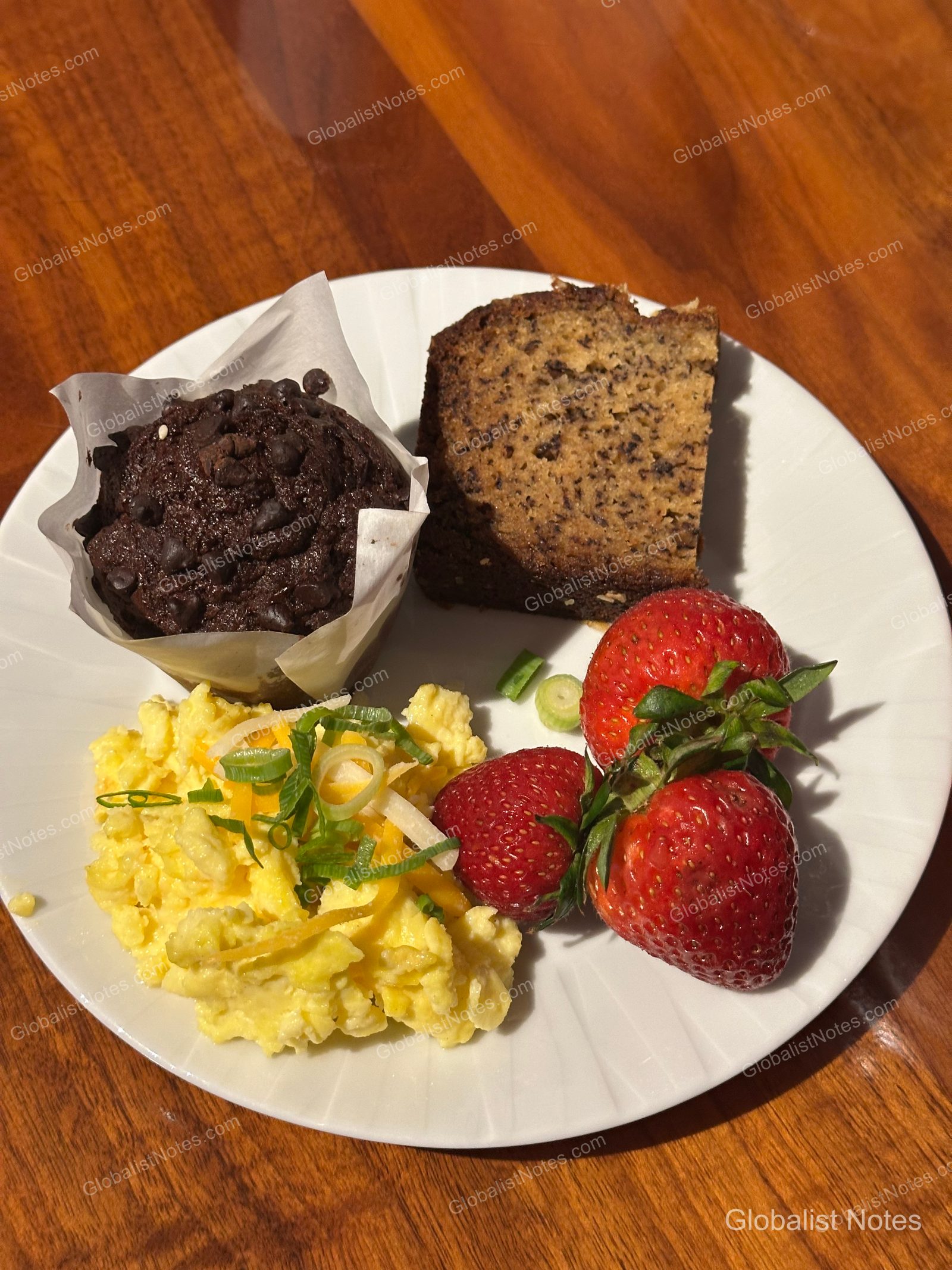 A breakfast plate featuring a chocolate muffin, a slice of banana bread, scrambled eggs, and three fresh strawberries.